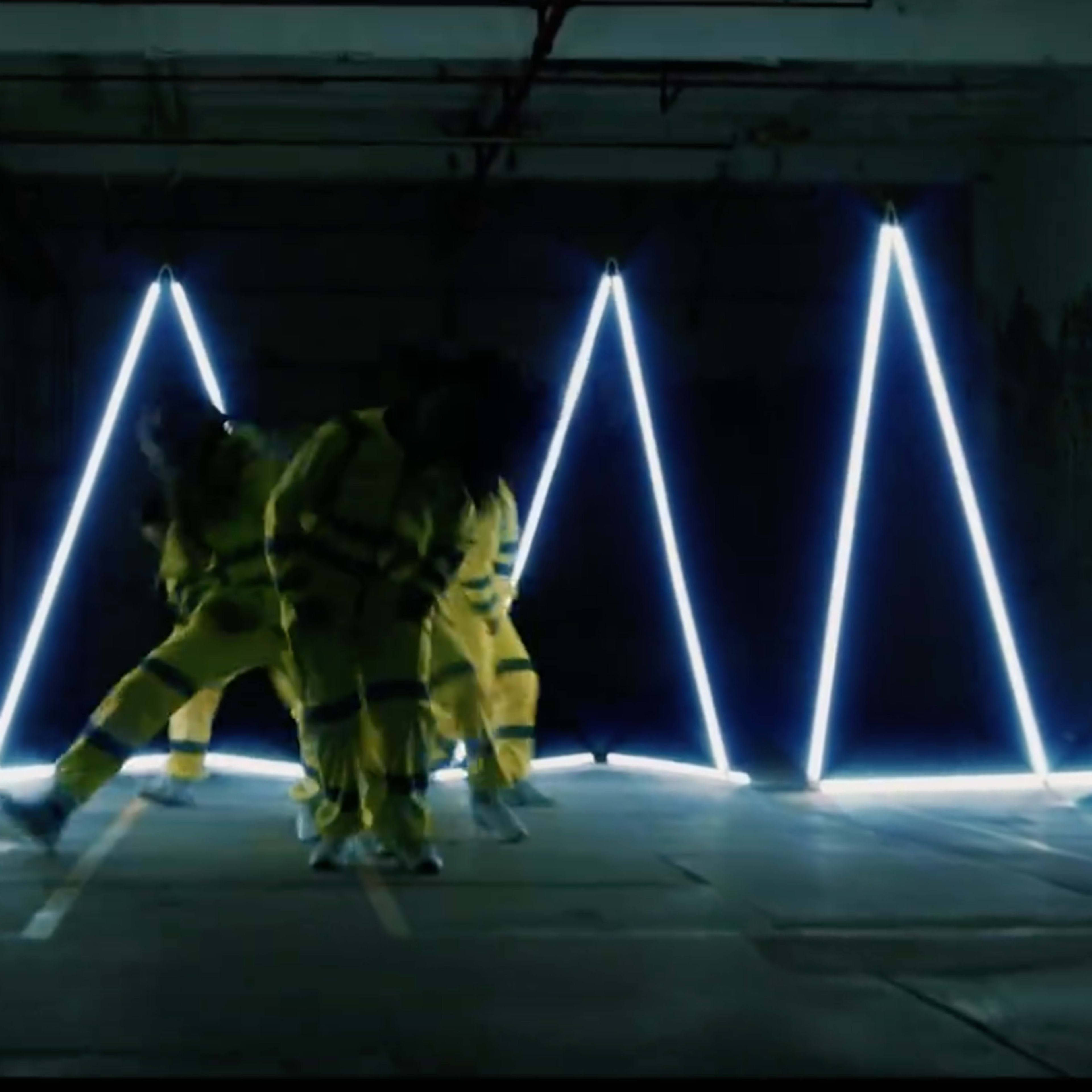 A group of dancers in yellow outfits performing in a dimly lit space with bright, angular LED lights creating a geometric pattern.