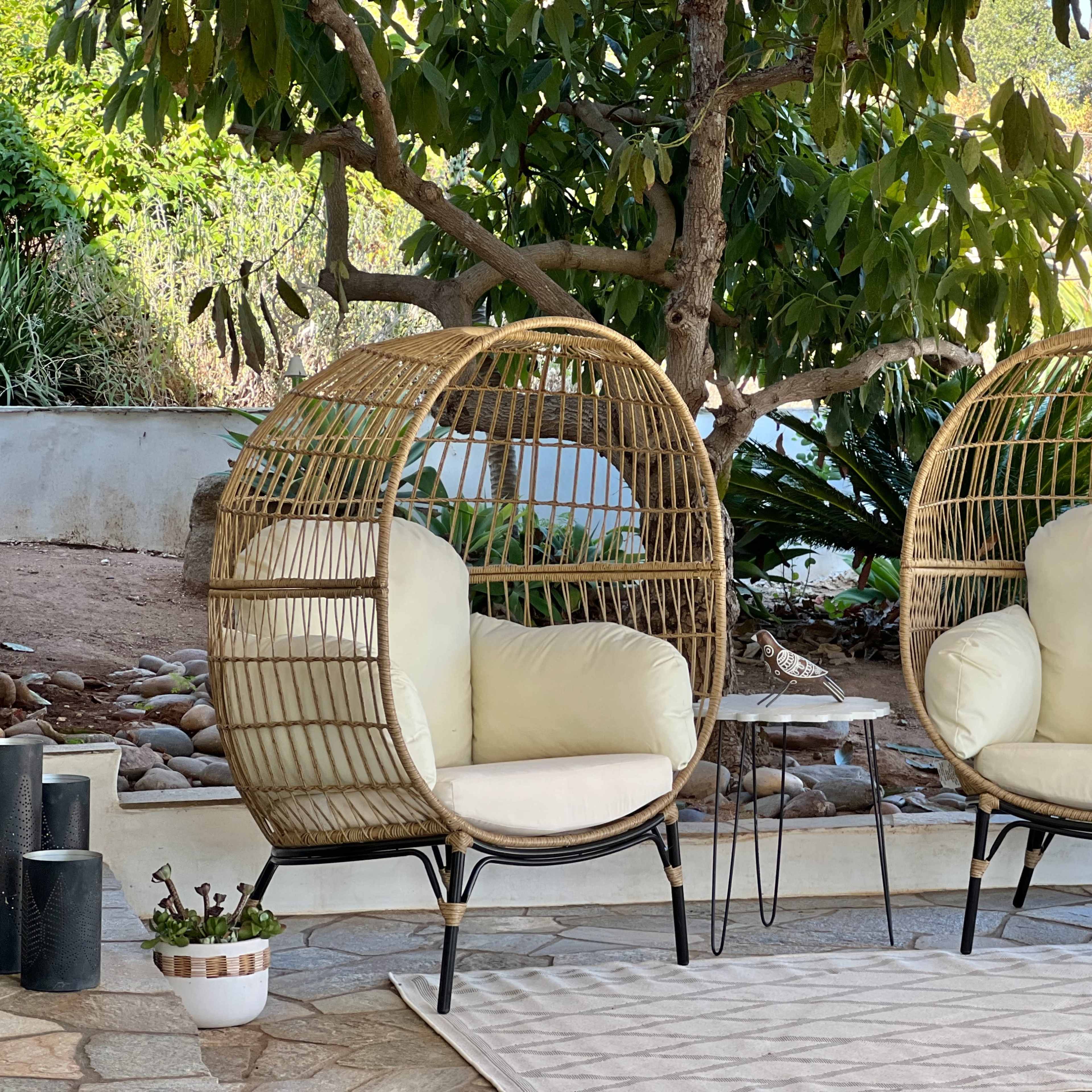 The scene features two circular rattan chairs with cushions placed next to a small side table, surrounded by greenery and stone landscaping.