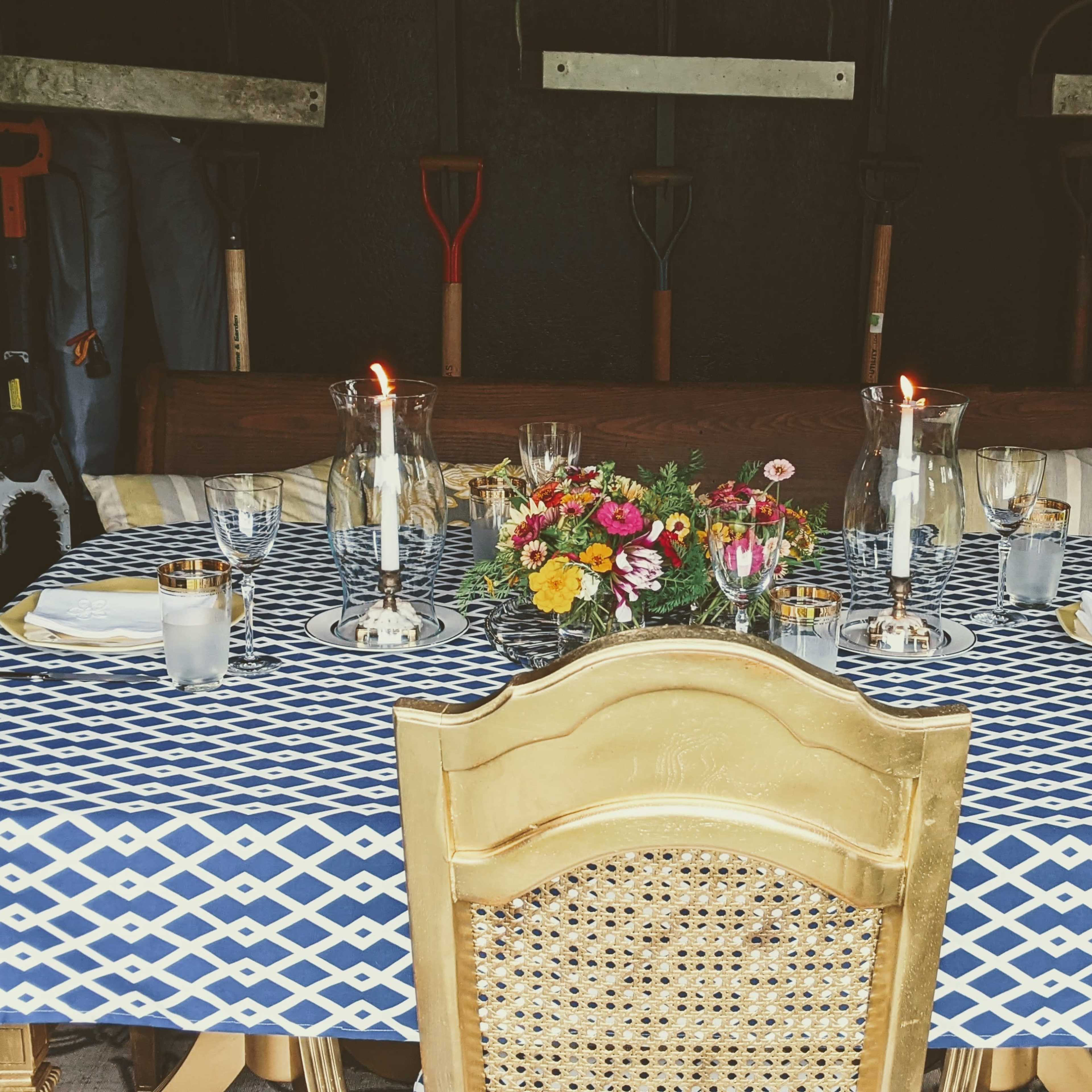 A dining table is set with a blue and white patterned tablecloth, candles, flowers, and glassware, surrounded by gardening tools hanging on the wall behind it.