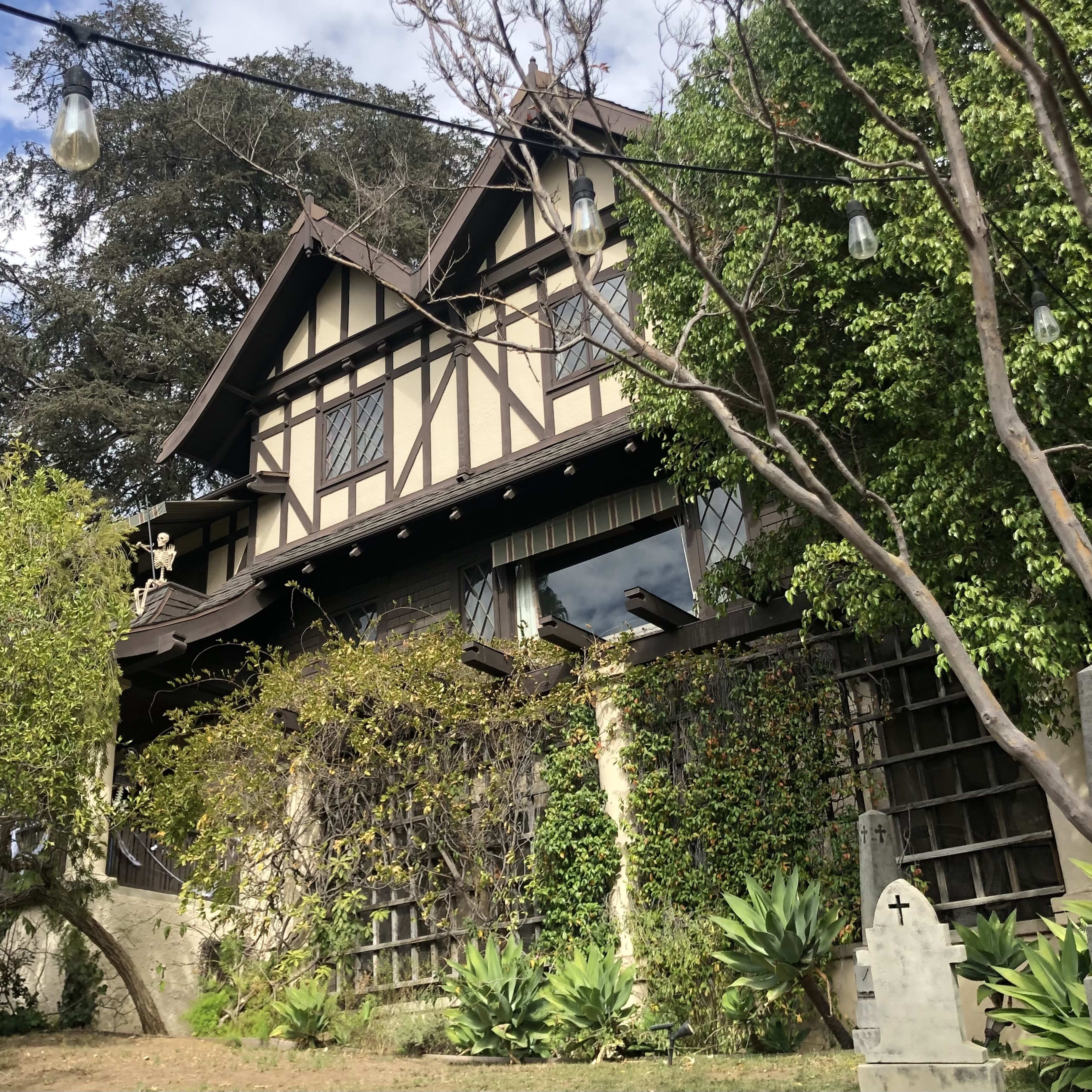 A large Tudor-style house with a stone exterior and a sloping roof is surrounded by greenery and decorative plants.