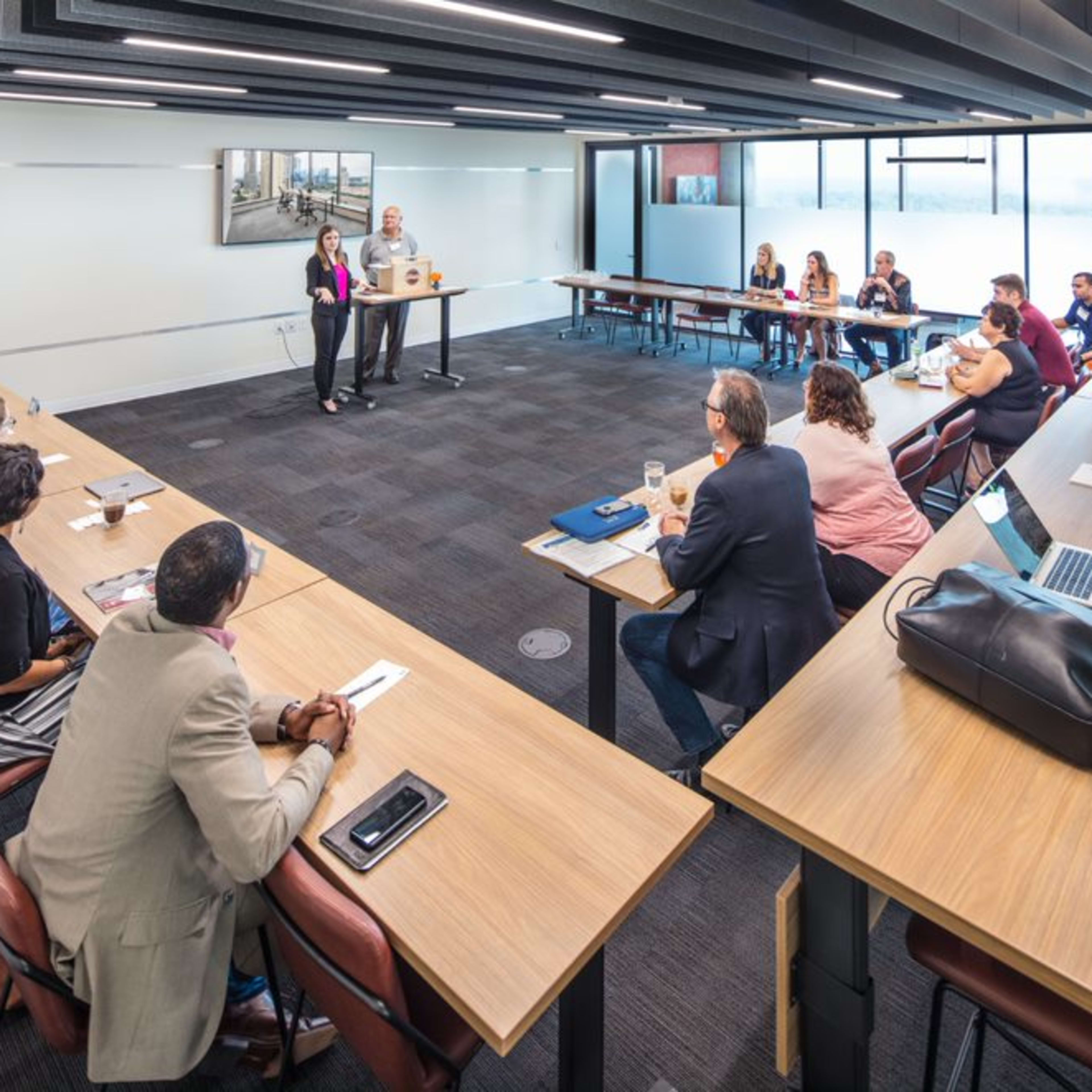 A group of people is seated at long tables in a conference room, while two speakers present in front of a large window.