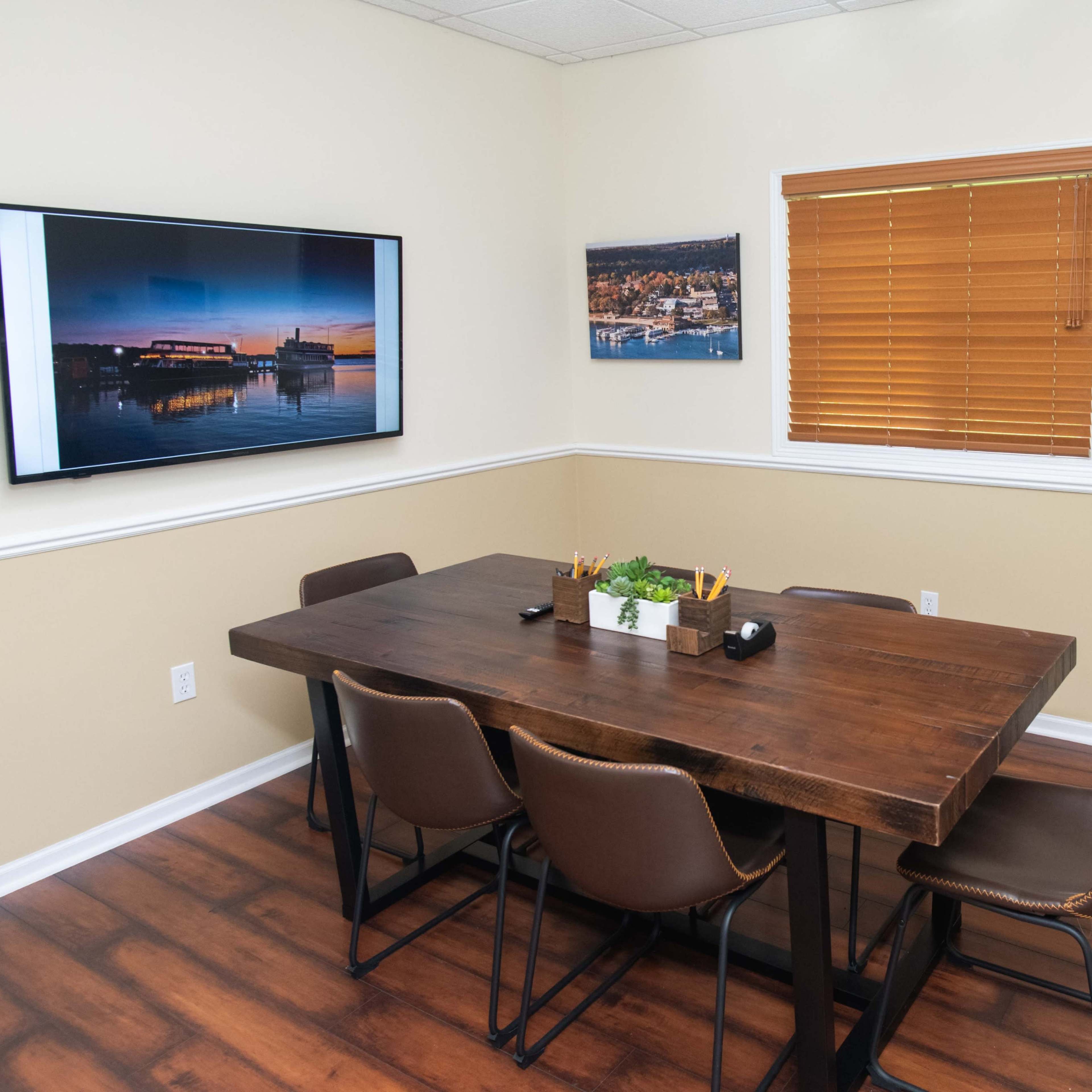 A conference room features a wooden table surrounded by chairs, with a television mounted on the wall and artwork displayed near a window with blinds.
