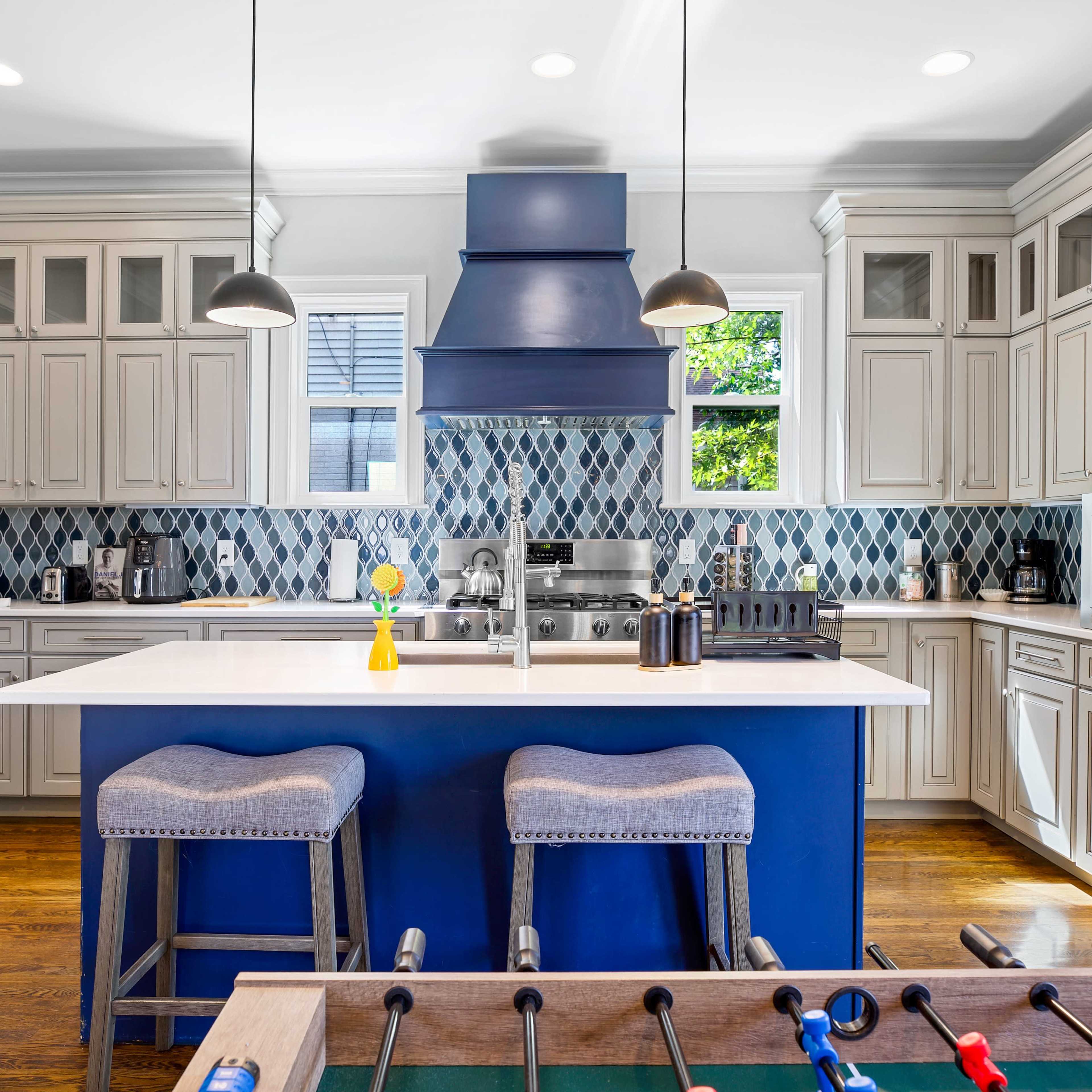 A modern kitchen features light gray cabinets, a blue accent chimney, and a large island with bar stools, alongside a foosball table in the foreground.