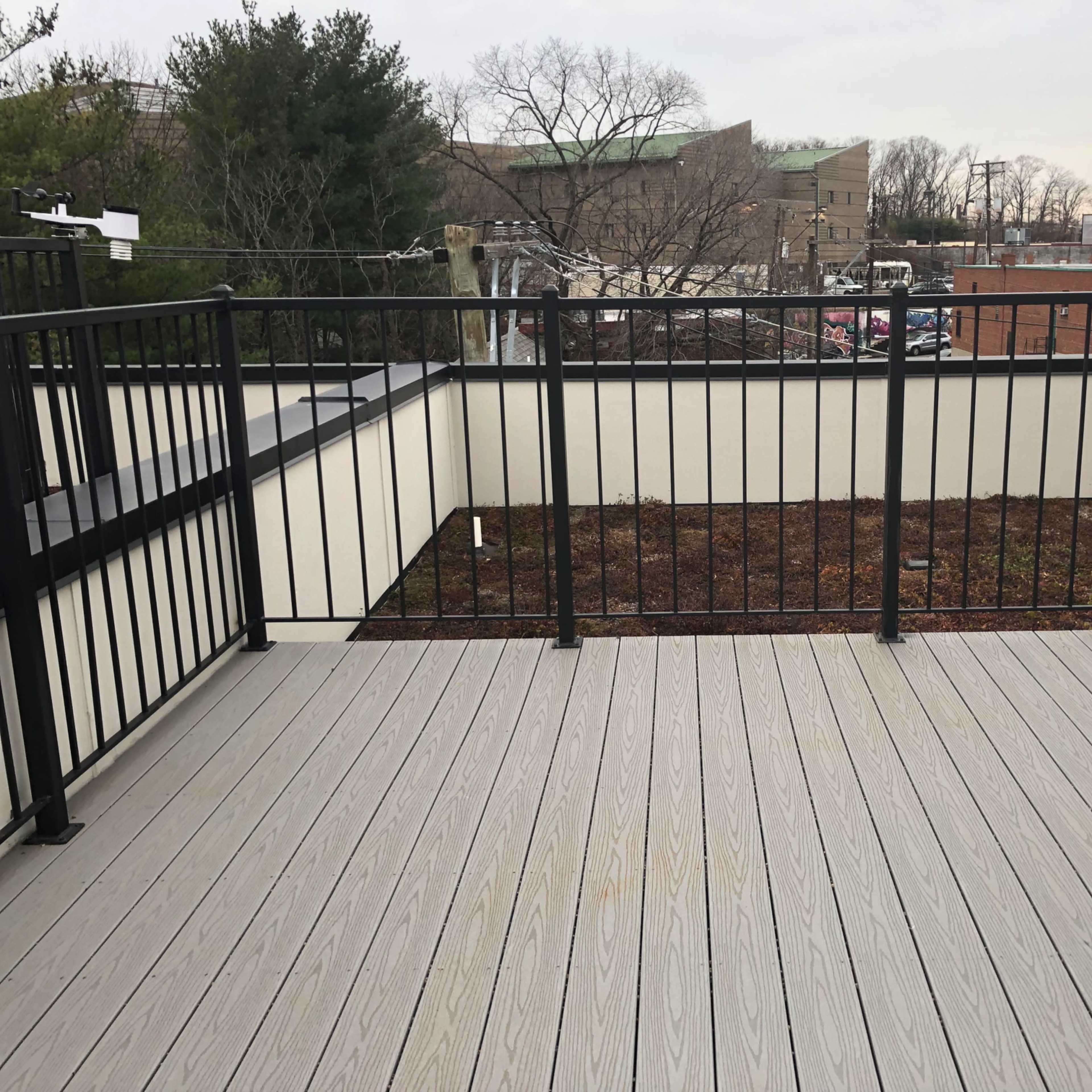 A rooftop terrace with a wooden floor and a metal railing surrounding a small, enclosed patch of bare soil and grass.