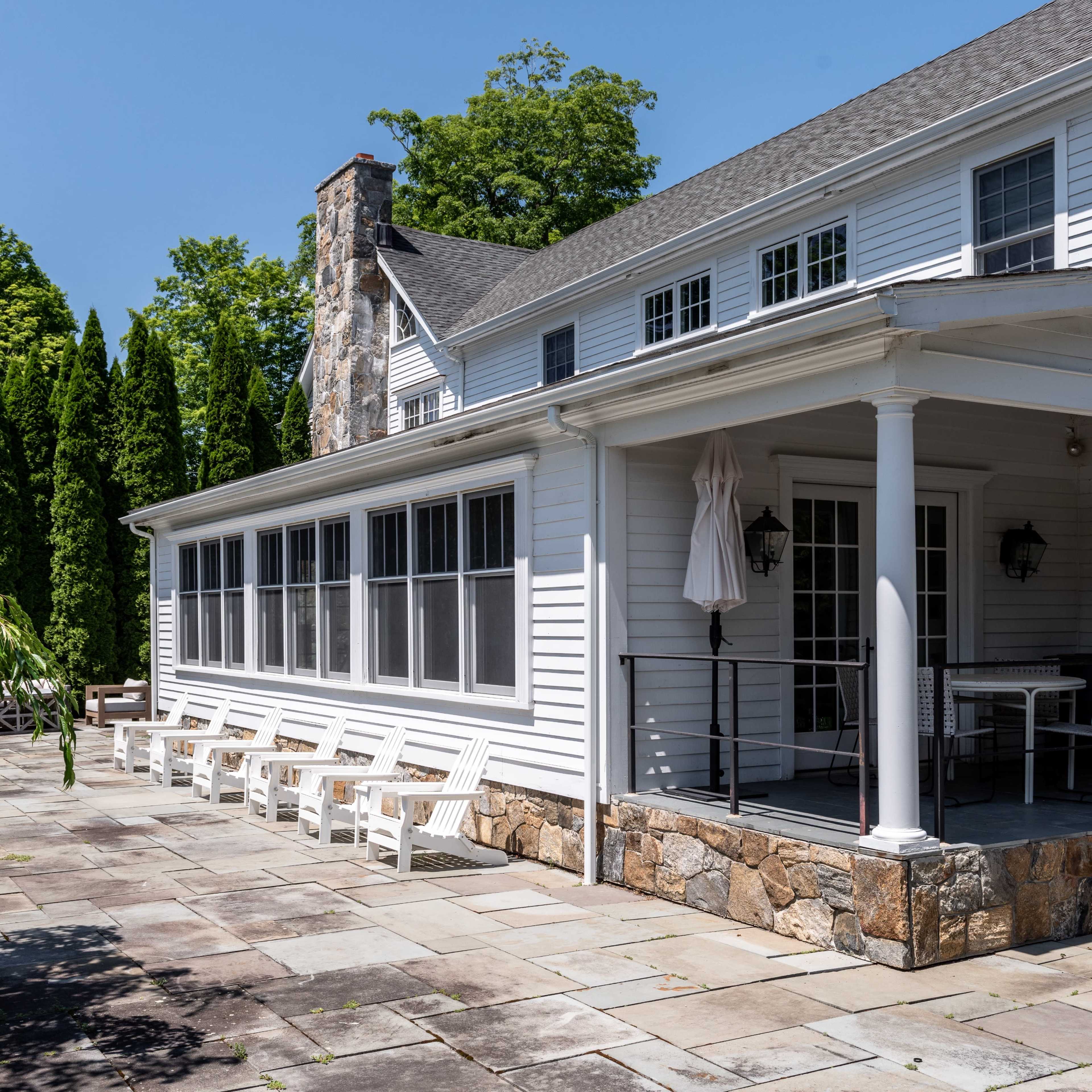 A two-story white house with a stone chimney features a screened porch and a stone patio surrounded by tall trees.