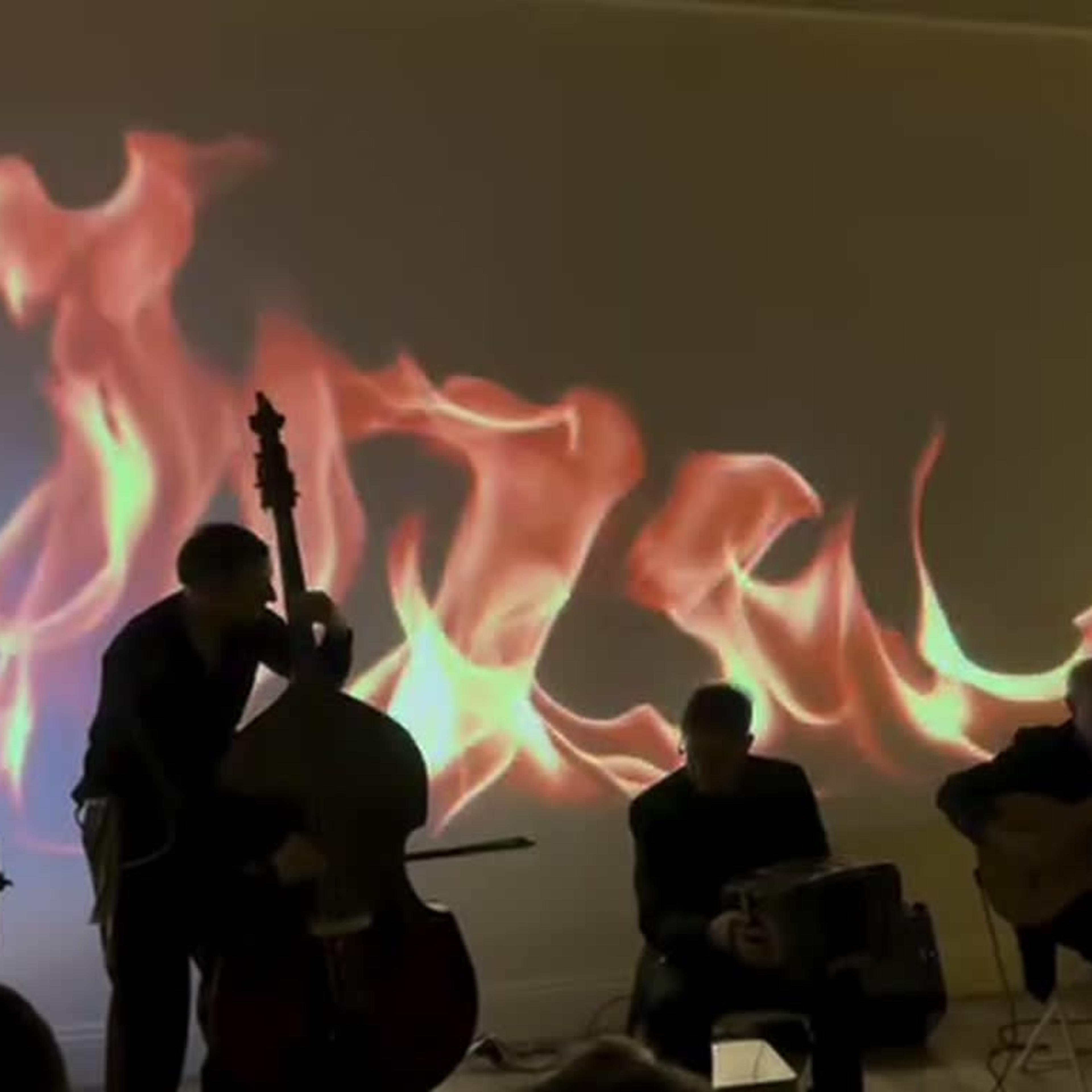 Musicians perform in front of a wall displaying a fiery projection.