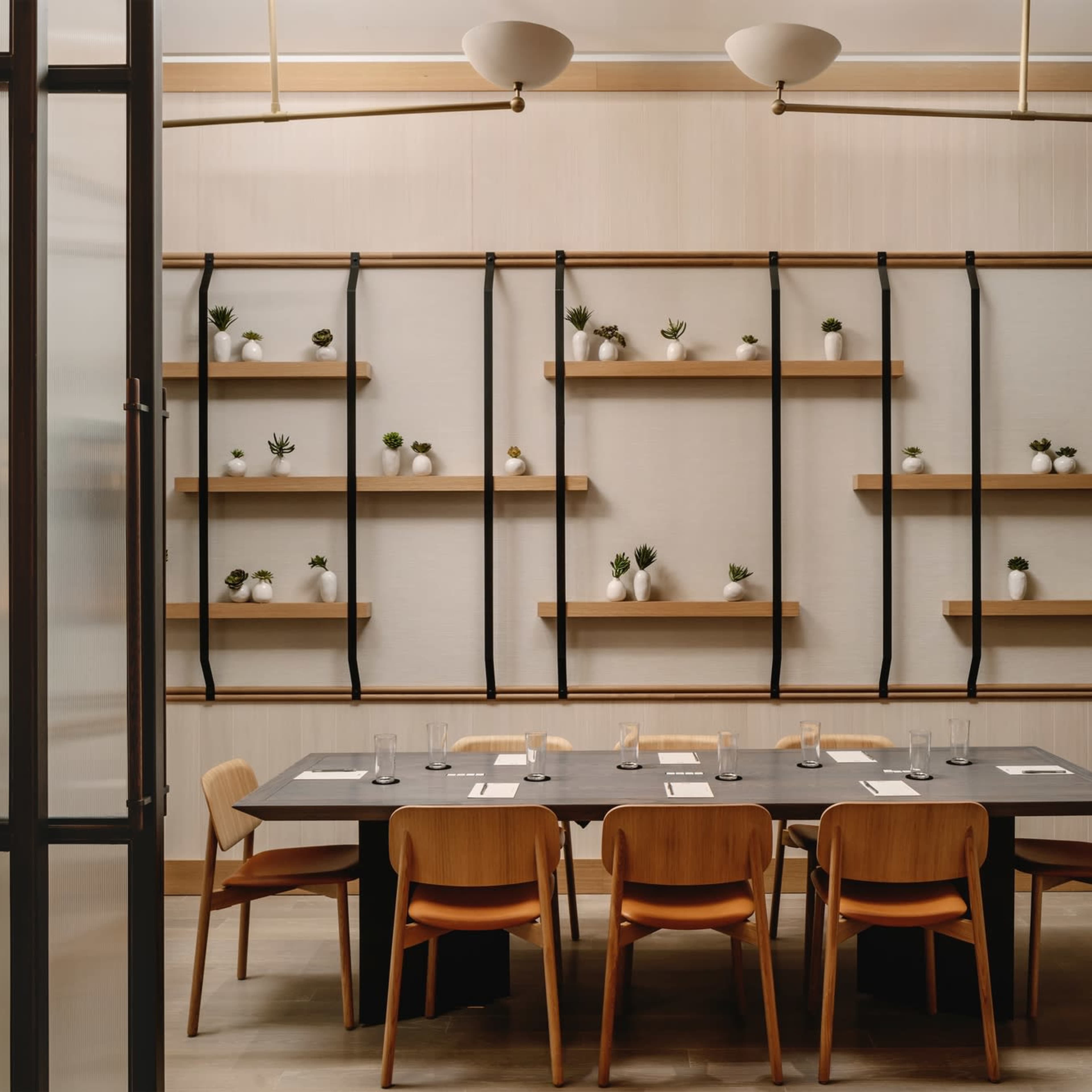 A dining room features a long table surrounded by wooden chairs, with wooden shelves displaying small plants on the wall.