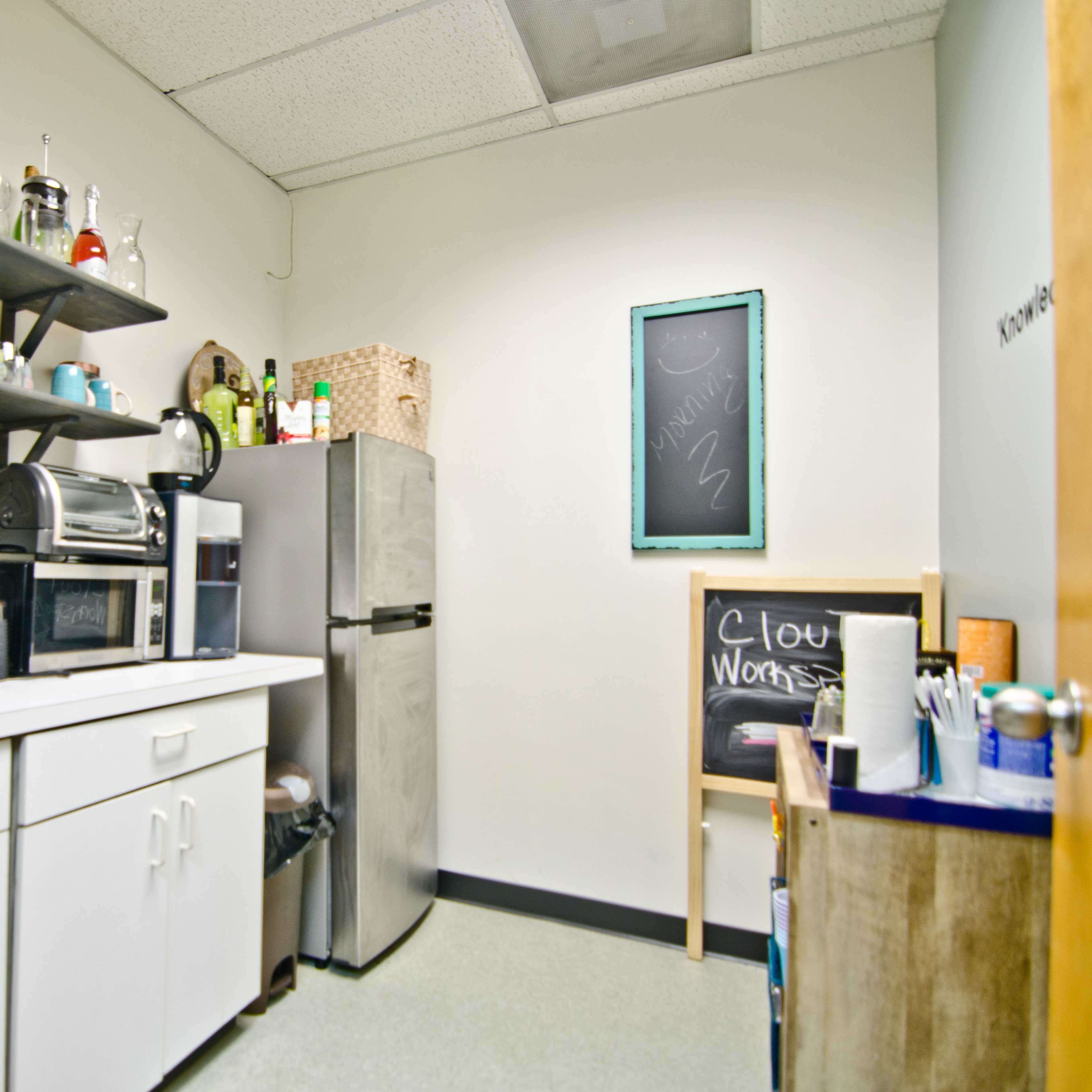 A small kitchen area with a sink, coffee machine, refrigerator, and shelves displaying various glassware and utensils.