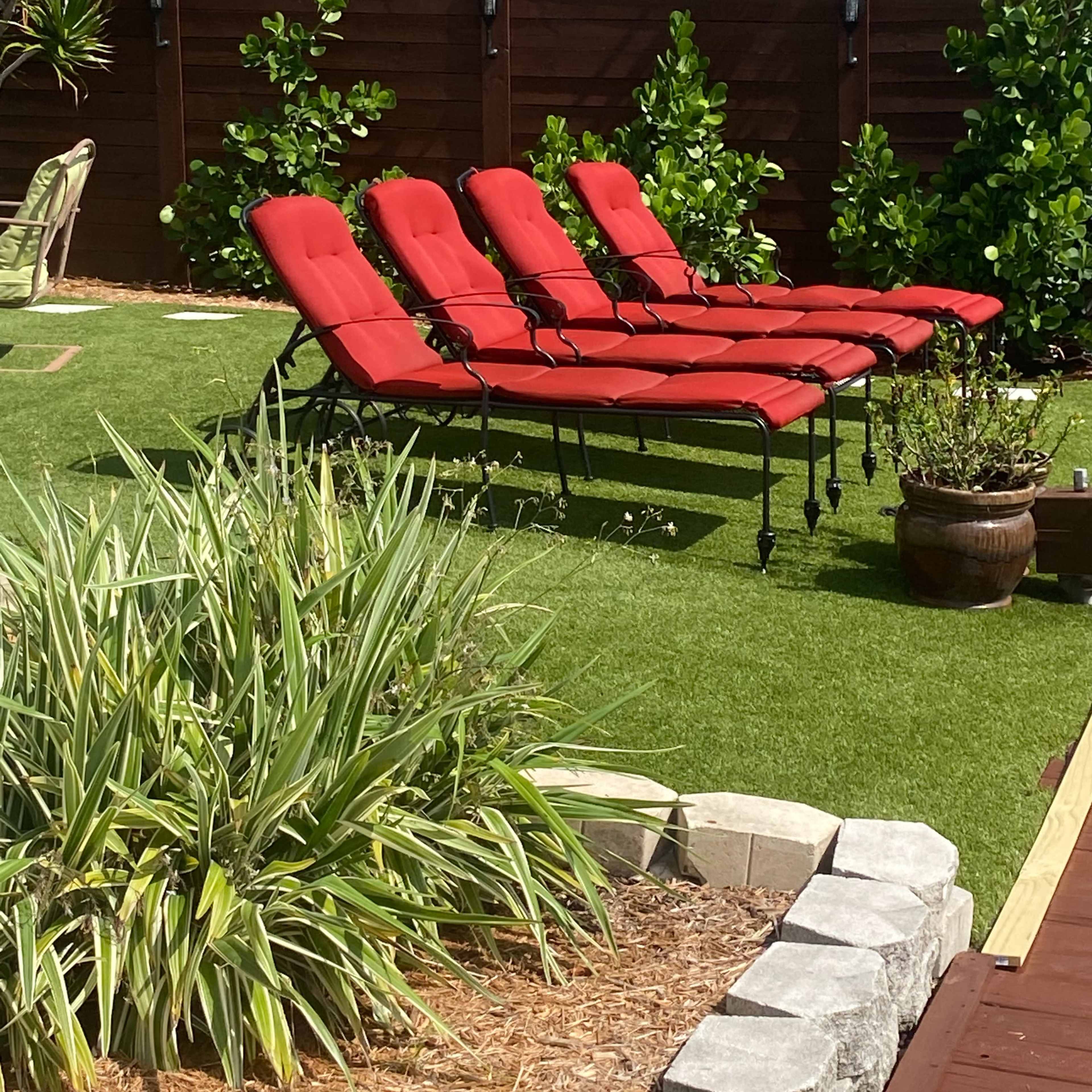 Four red lounge chairs are arranged on a patch of green grass in a garden setting, surrounded by landscaping and wooden fencing.