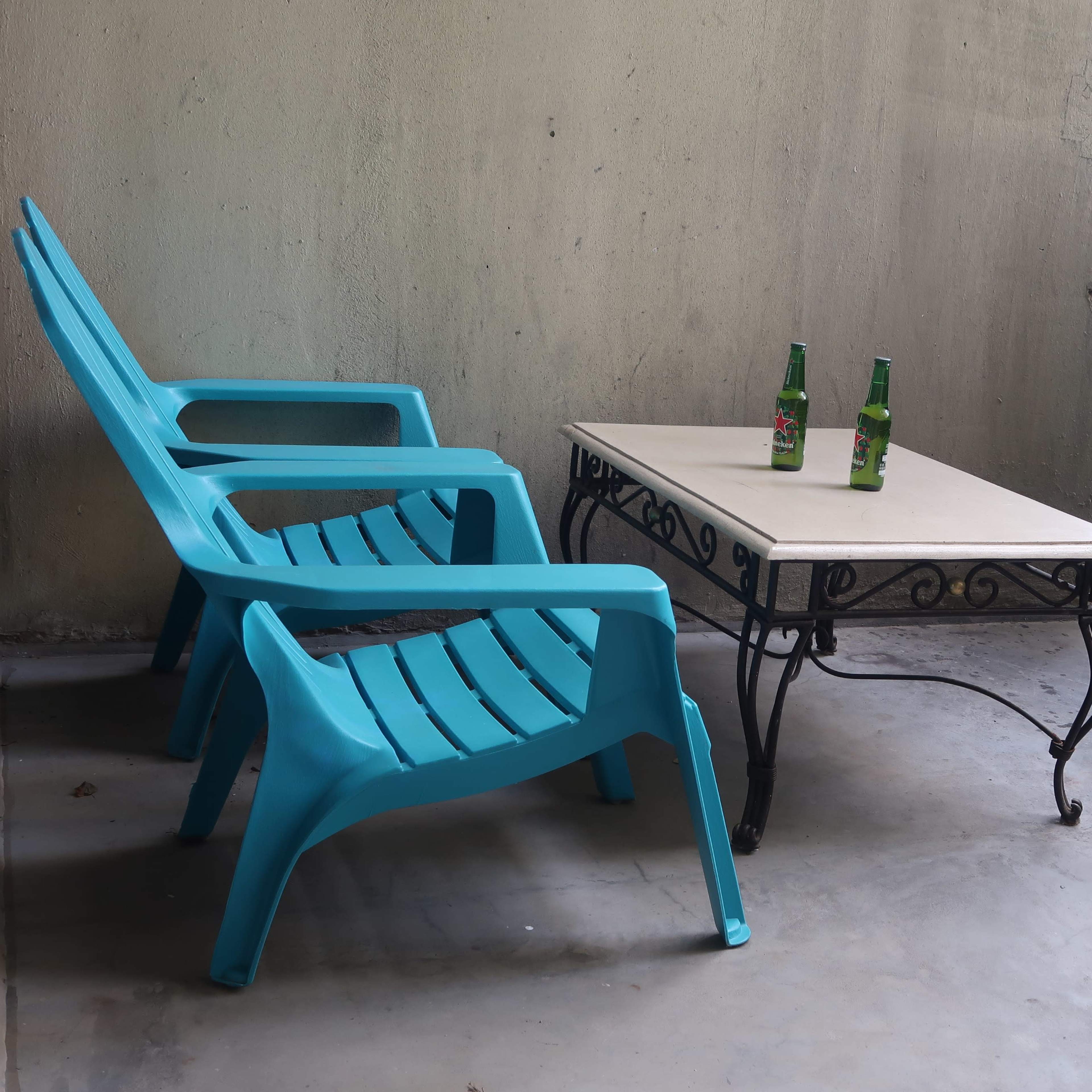 A small balcony features two turquoise plastic chairs and a metal table, with two green bottles placed on top.