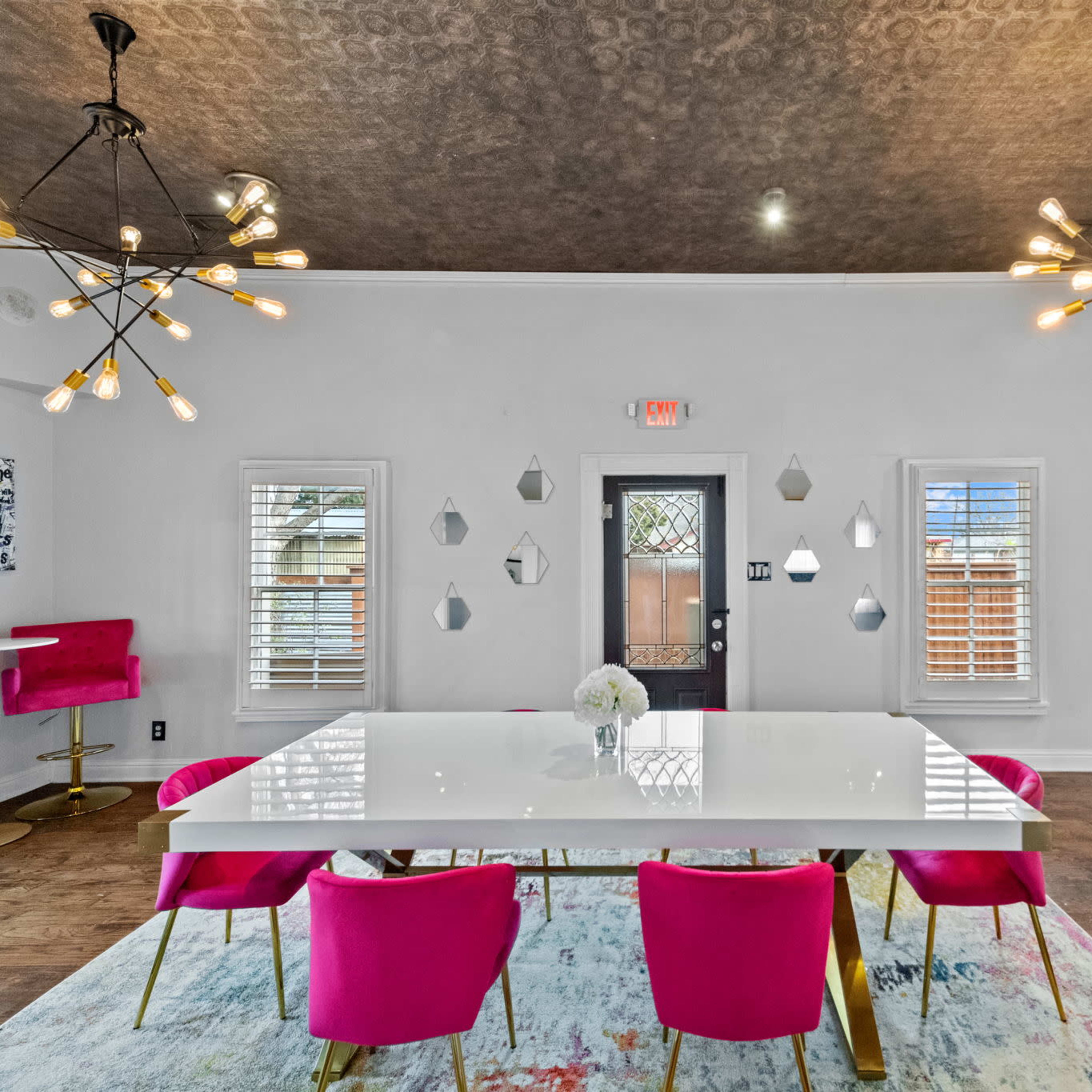 A modern dining area featuring a large white table surrounded by pink chairs, two unique light fixtures hanging from the ceiling, and decorative wall mirrors.