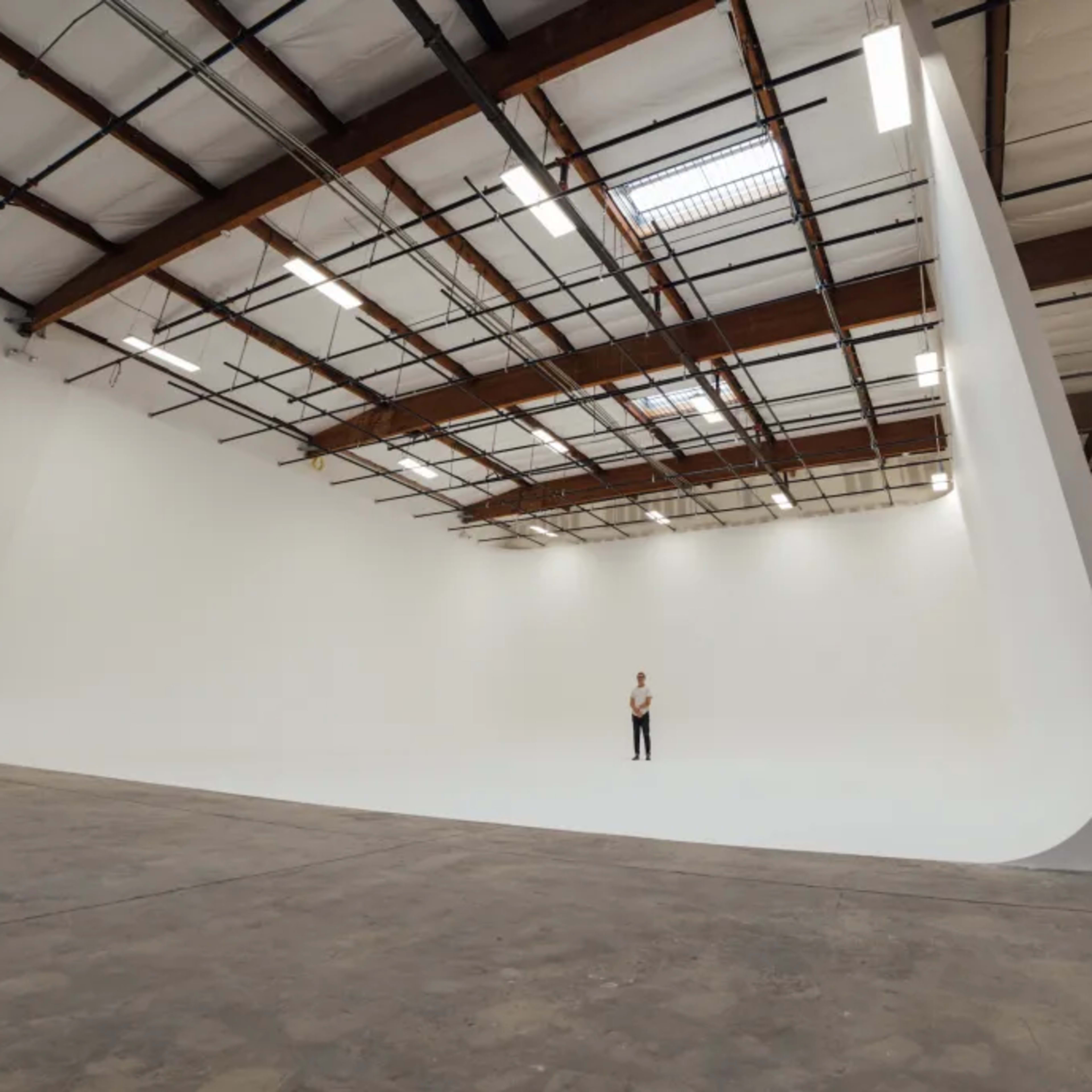 A person stands in a large, minimalist studio with a curved white backdrop and exposed wooden beams on the ceiling.