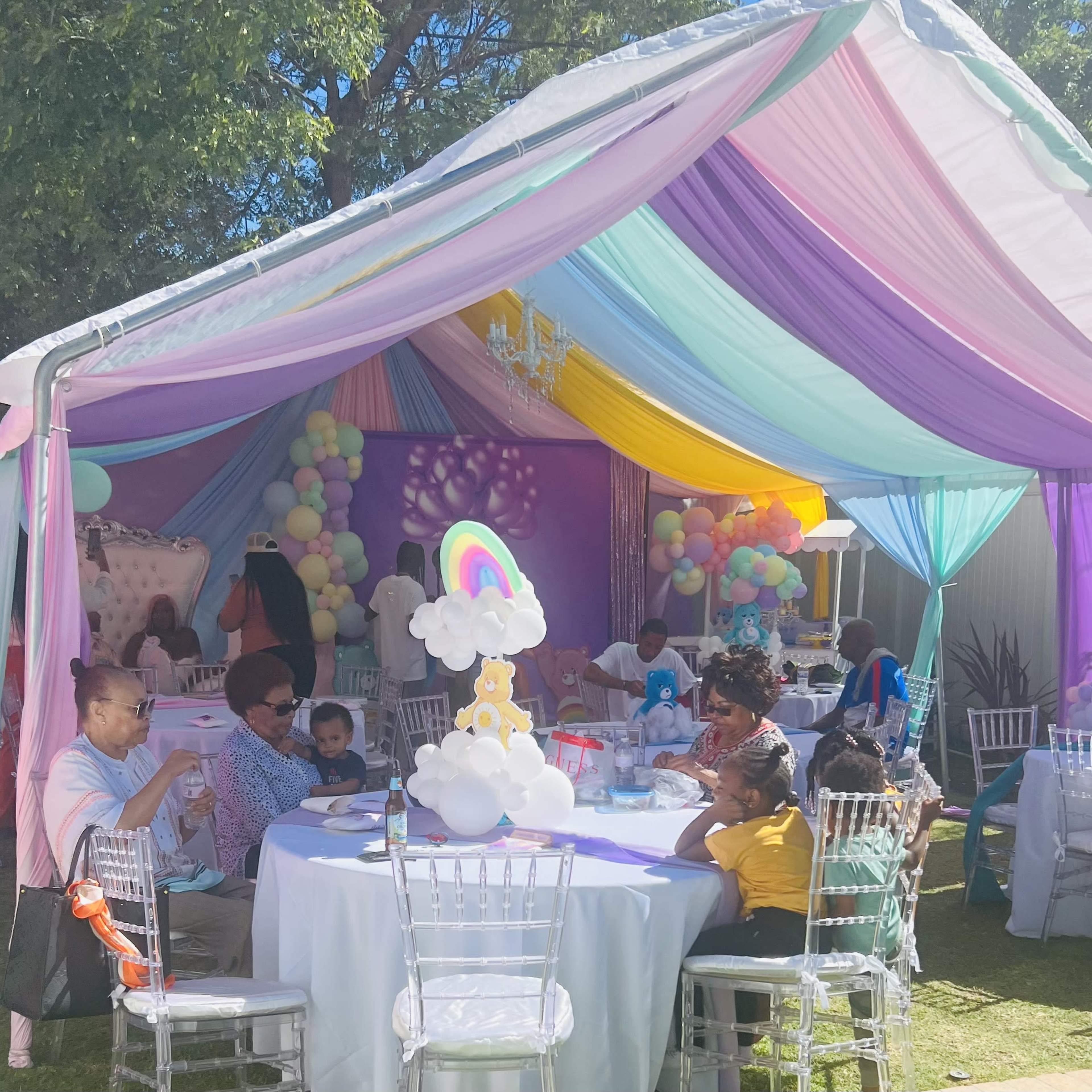 A colorful tent is set up outdoors with guests seated at tables adorned with decorations, enjoying a celebration.