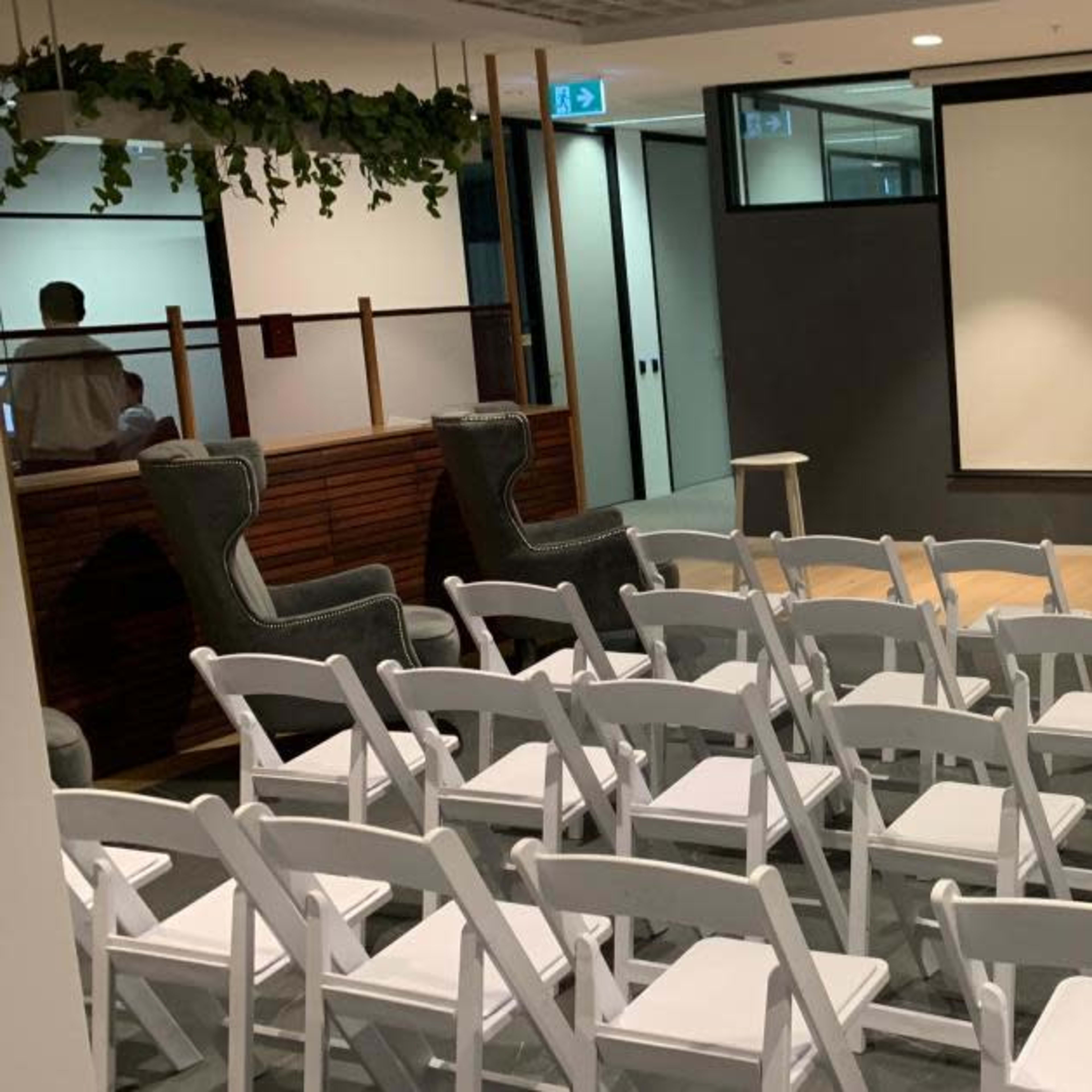 A small auditorium setup with white chairs arranged in rows facing a large blank screen, accompanied by three wooden stools on a stage area.