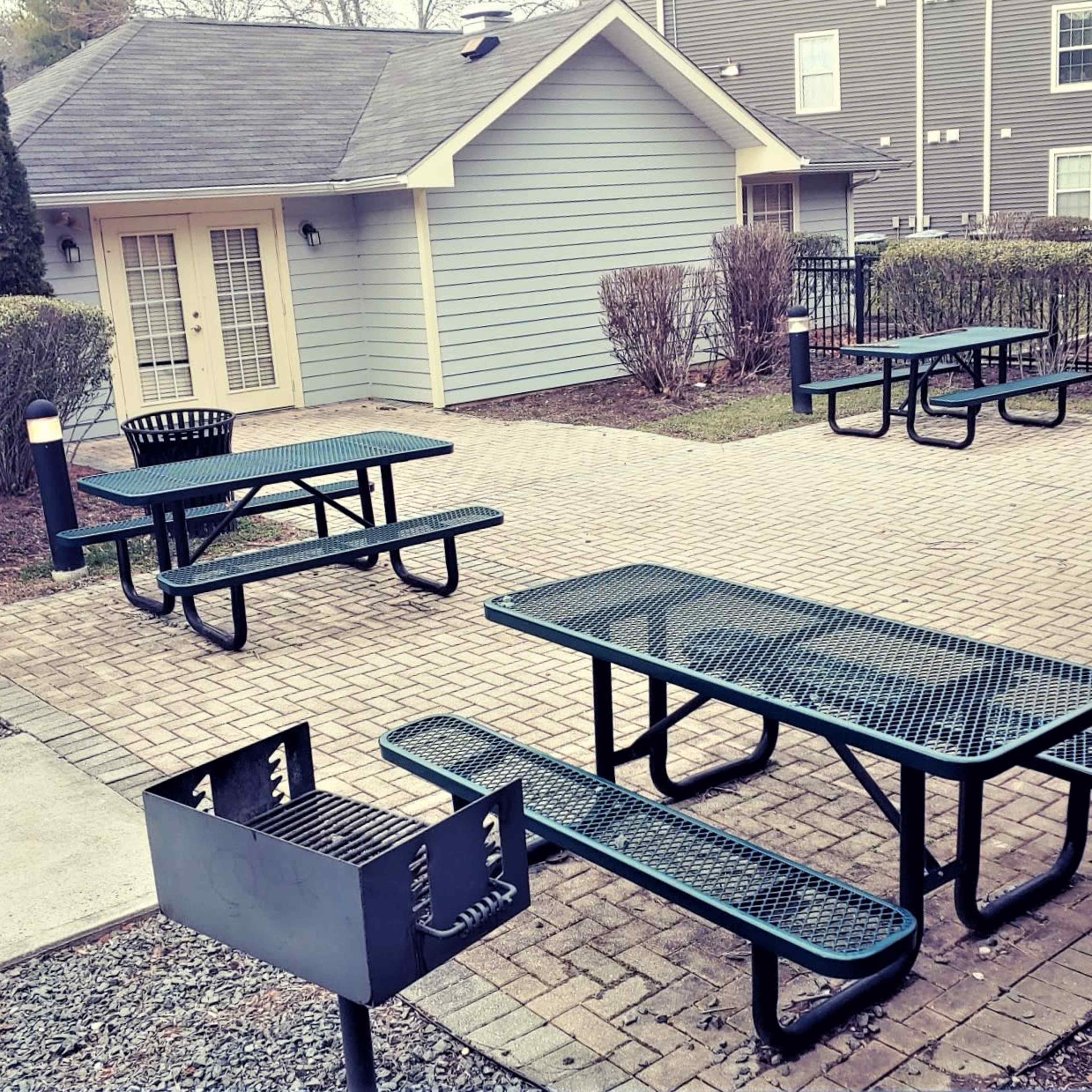 A patio area with several green picnic tables and grills surrounded by low shrubs and a gray building in the background.