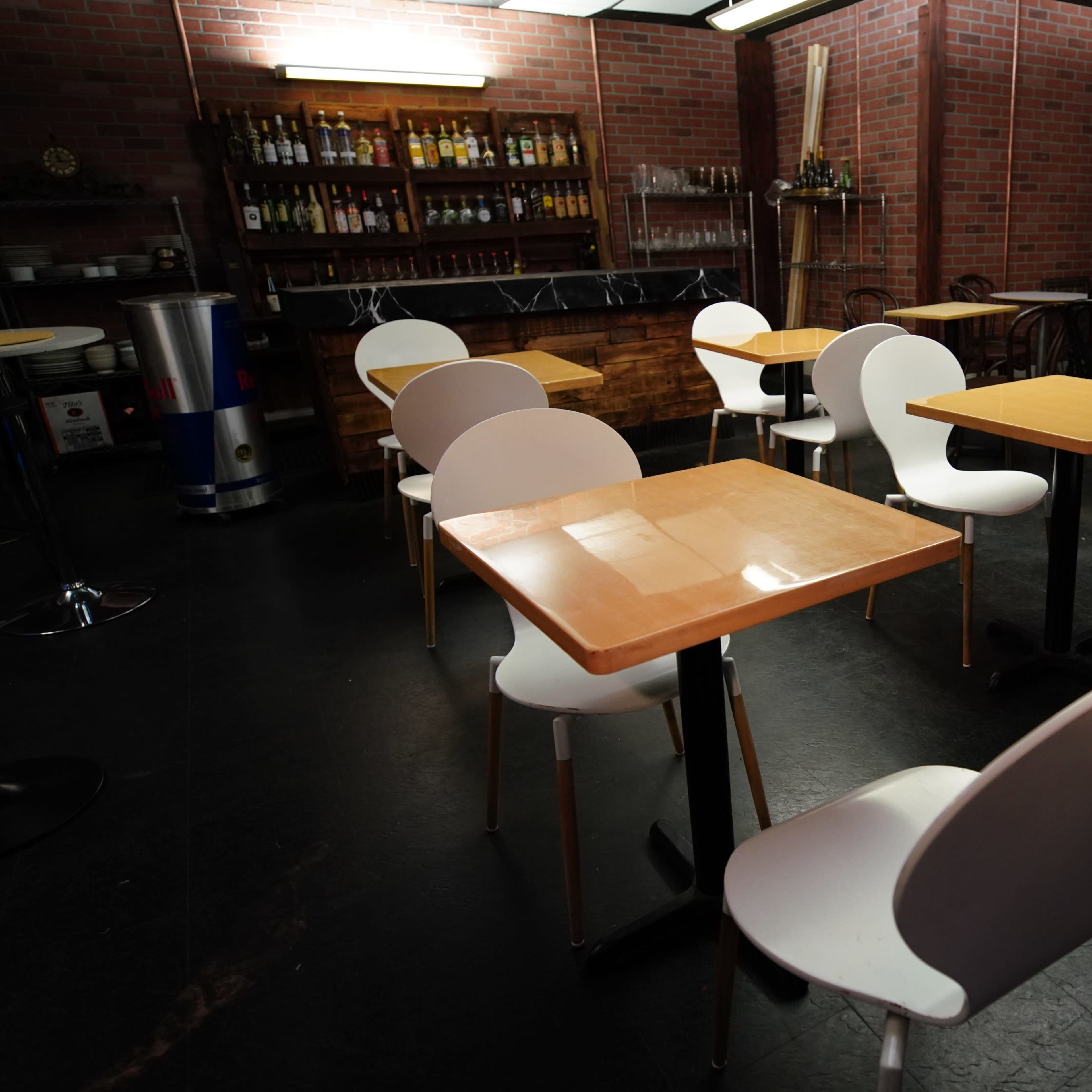 An empty restaurant interior with several tables and chairs arranged on a dark floor, surrounded by brick walls and a bar area in the background.