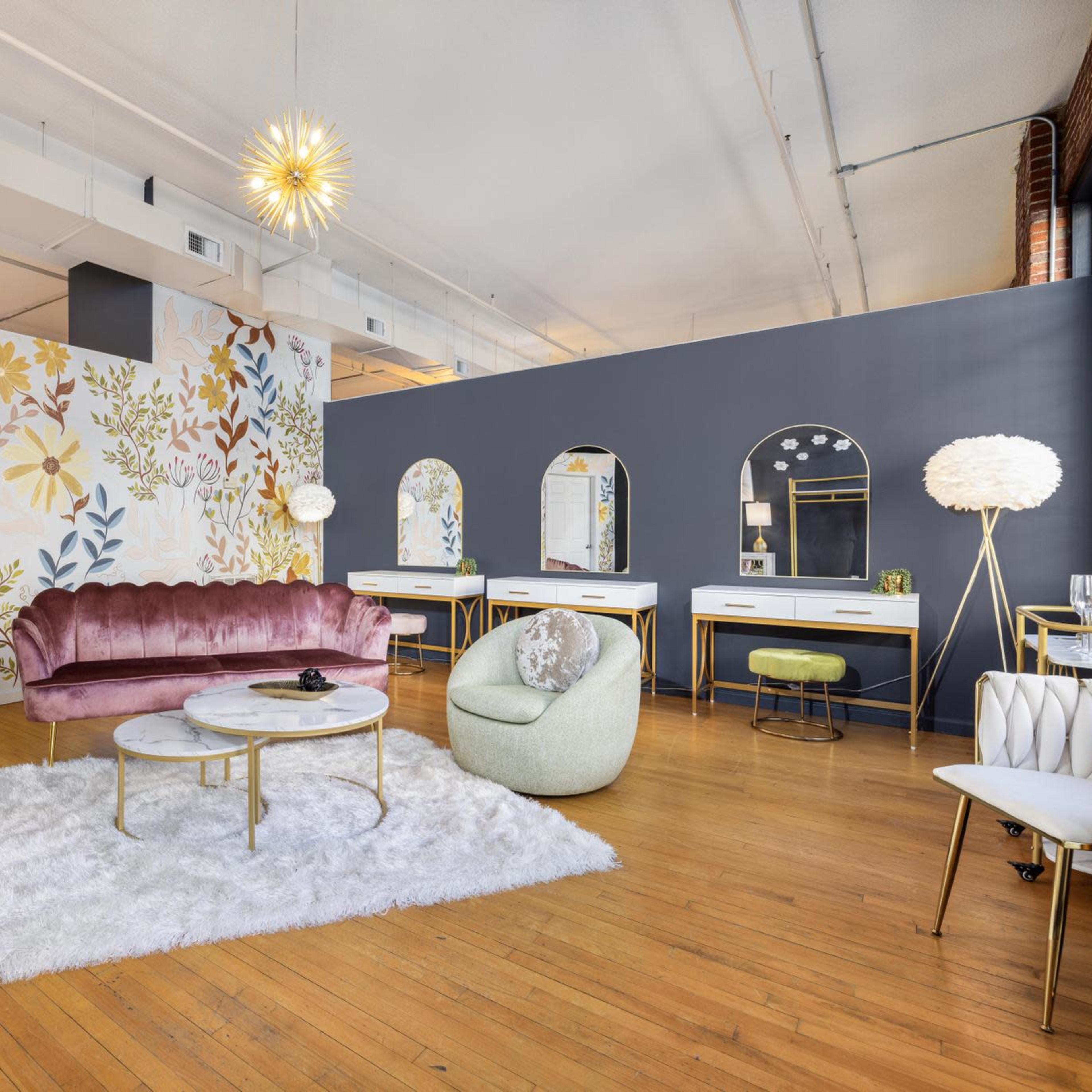 The room features a blend of floral wallpaper, a pink velvet sofa, two light-colored chairs, a round coffee table, and several mirrors set against a dark wall.