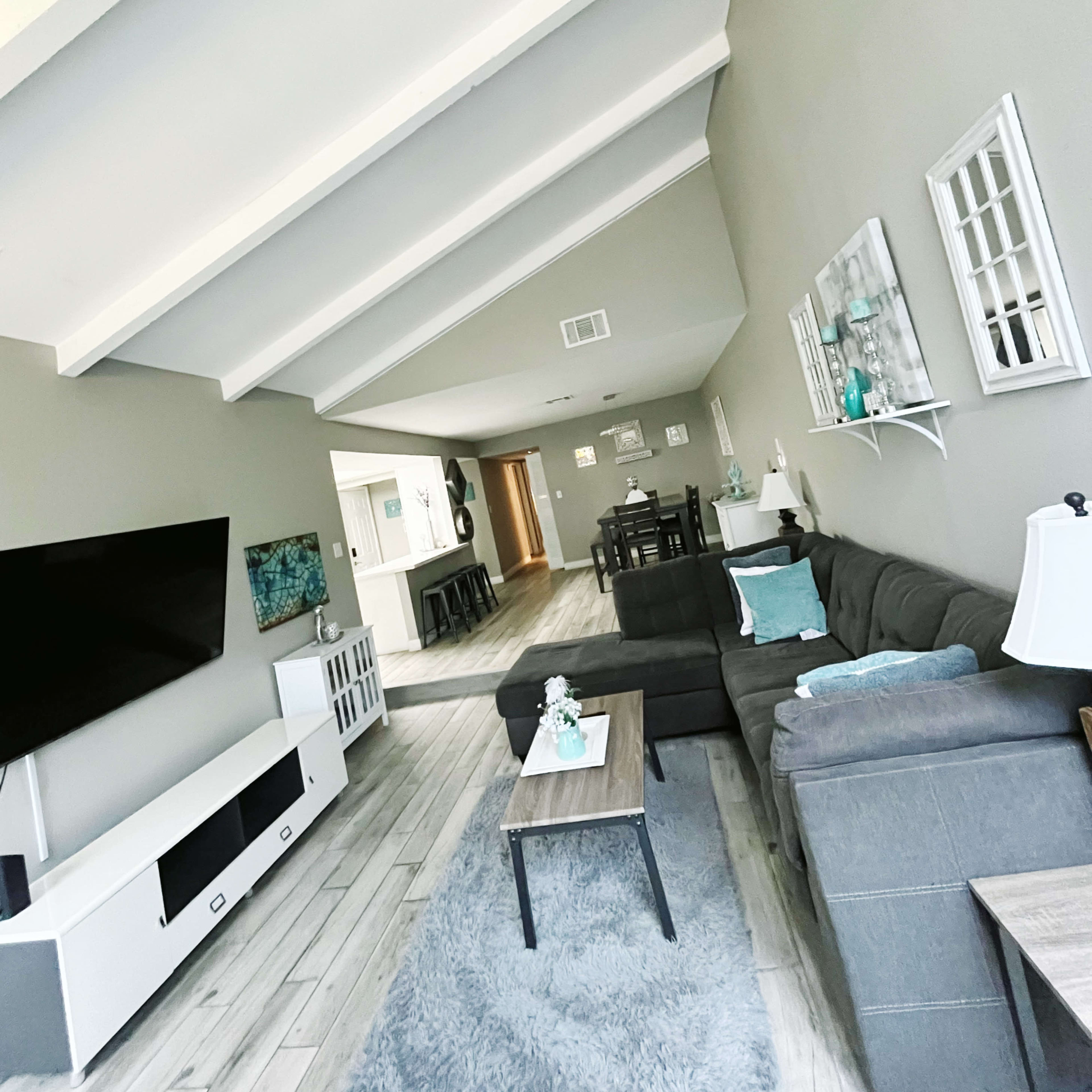 A spacious living room with a gray sectional sofa, a coffee table, and a television on a white media console, featuring vaulted ceilings and light wooden flooring.