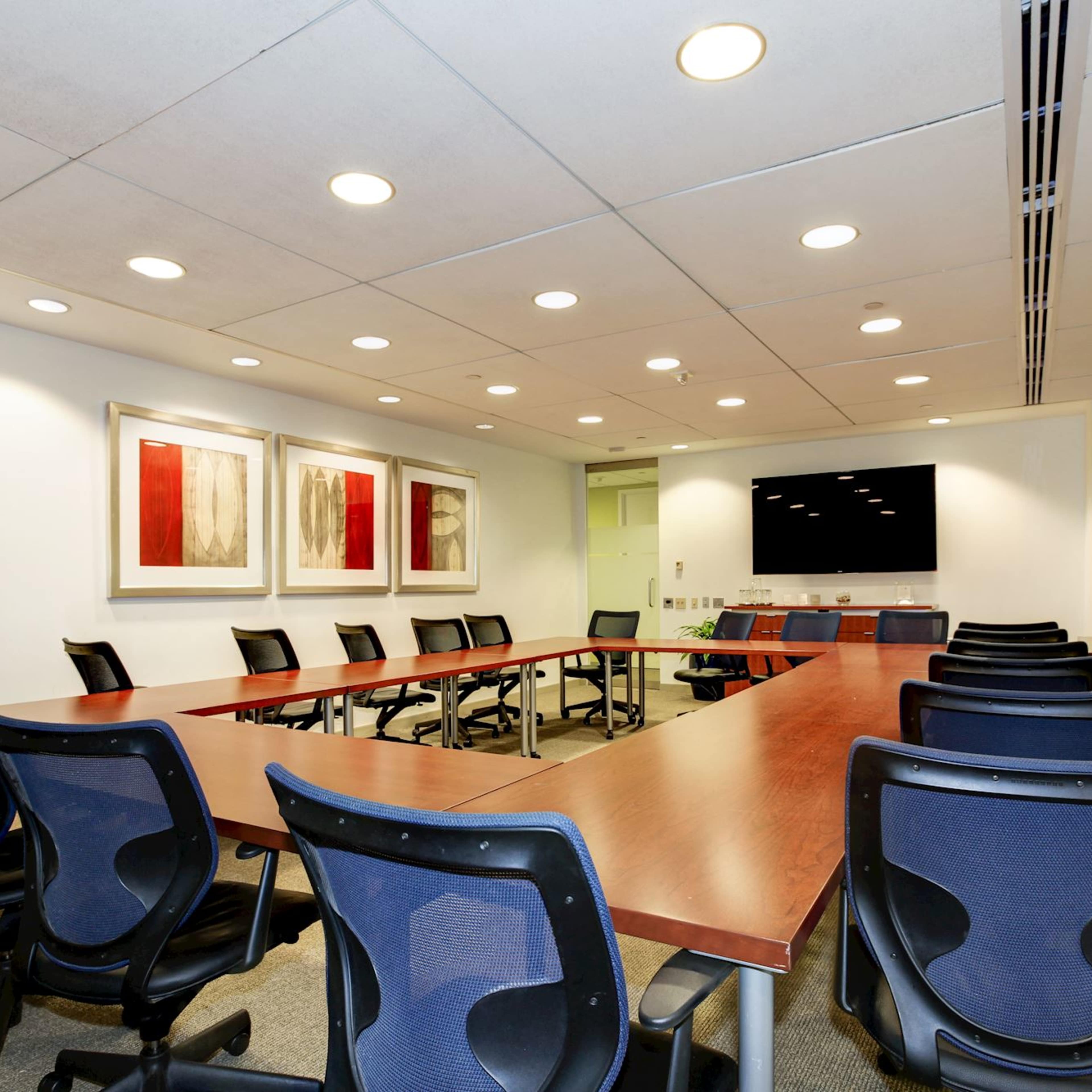 A modern conference room with a long table surrounded by black chairs, featuring two abstract paintings on the wall and a large screen at one end.