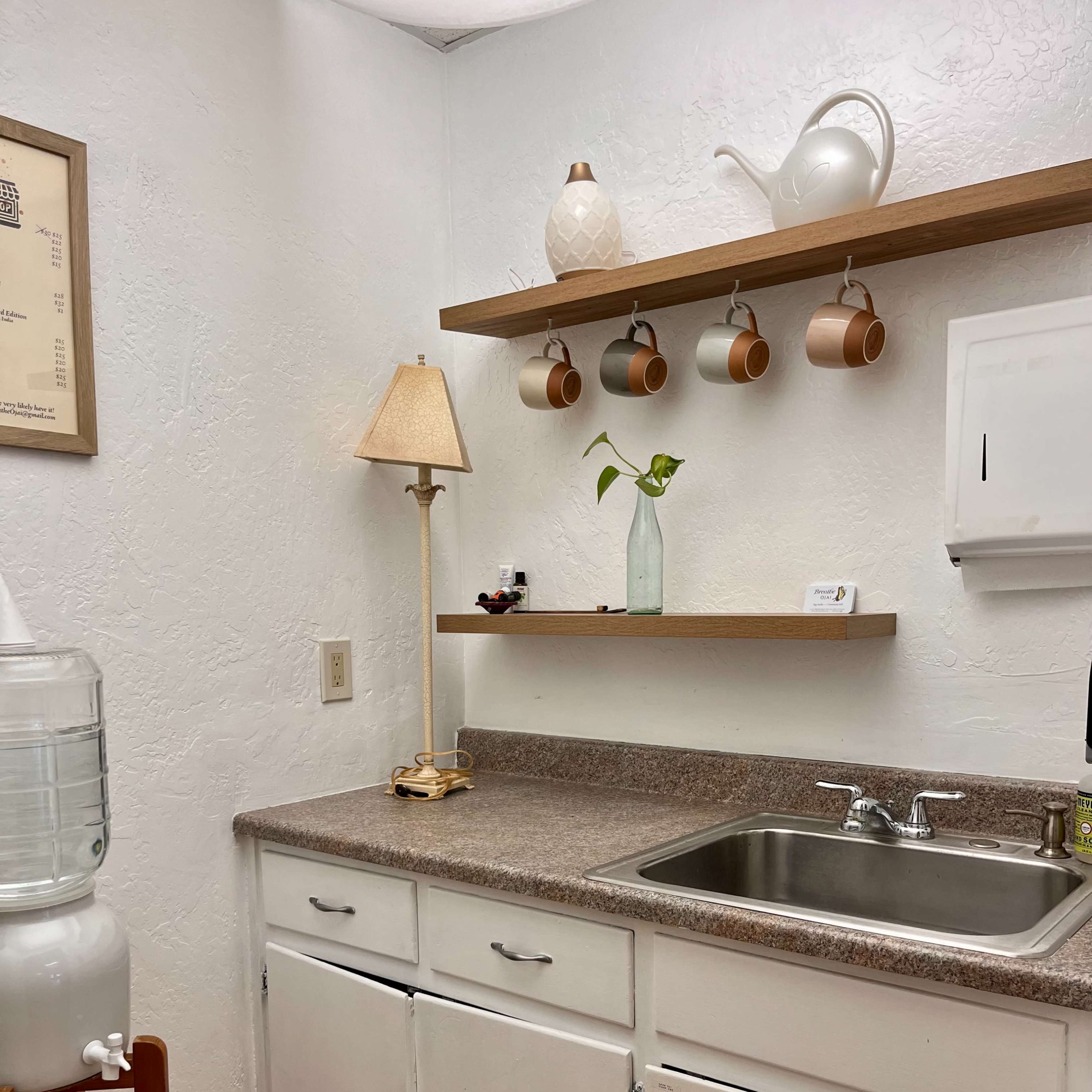 A small kitchen corner with a sink, a water cooler, a lamp, a shelf holding decorative items, and several hanging coffee mugs.