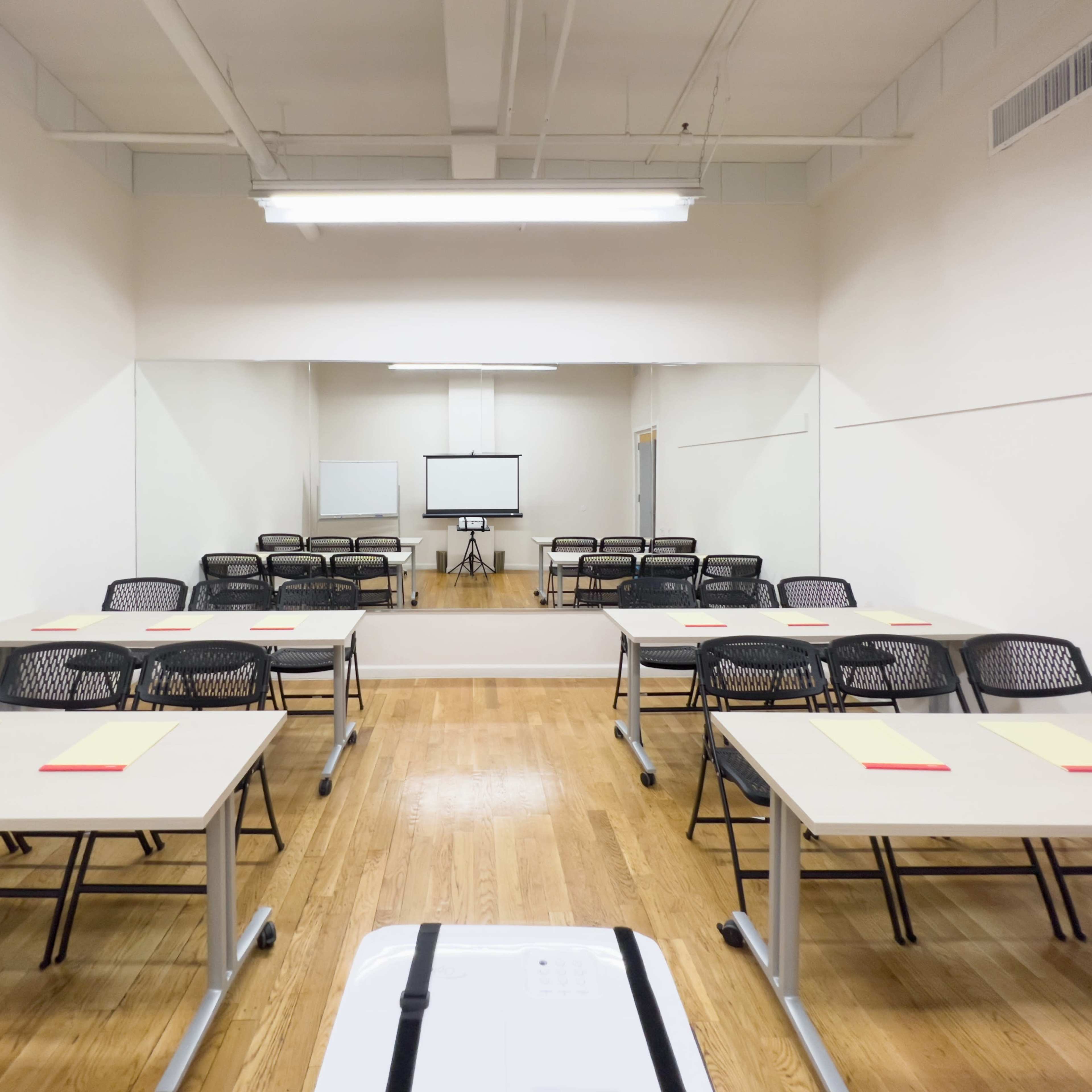 A classroom setting features multiple tables with chairs arranged facing a screen and a large mirror on one wall.