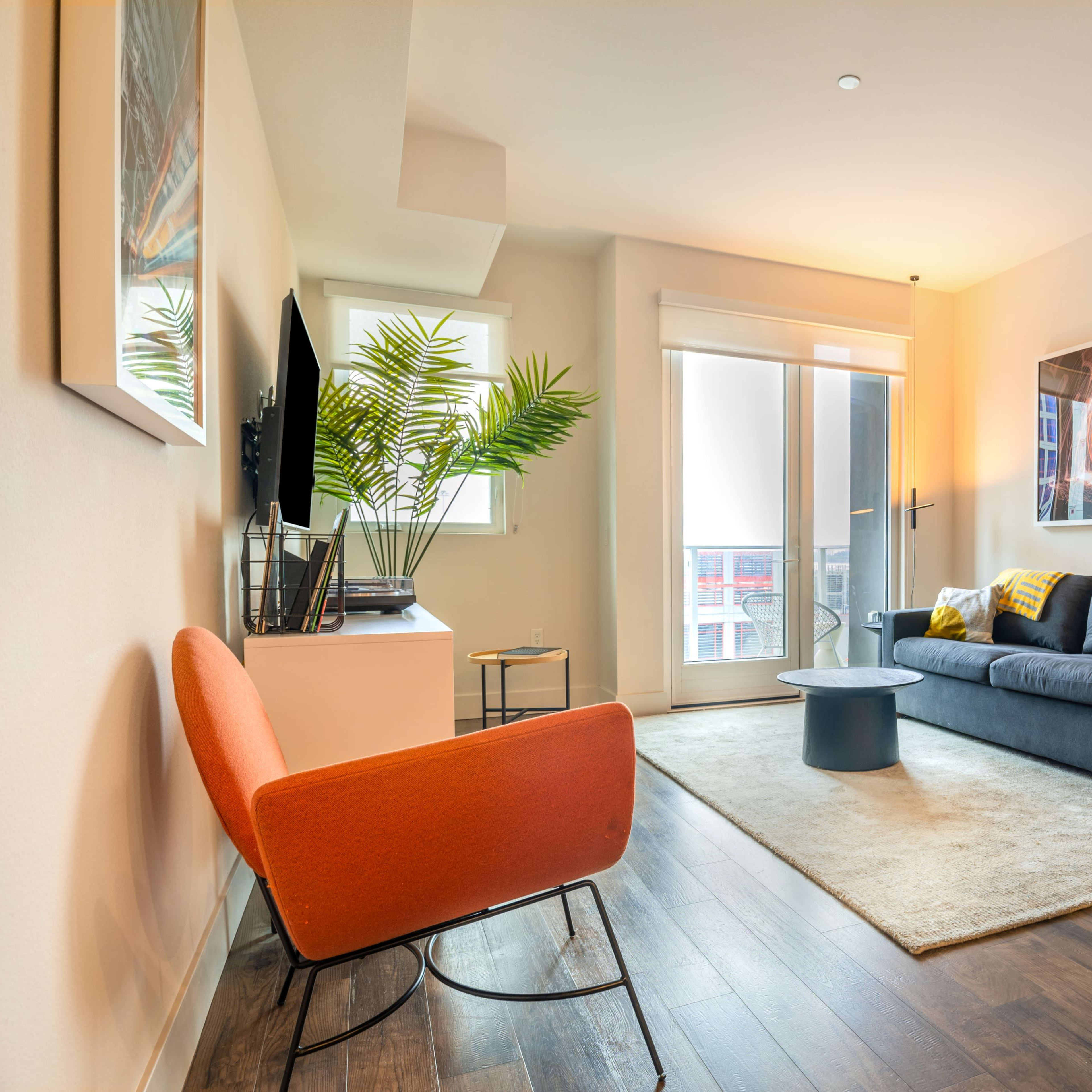 A modern living room features a gray sofa, orange chair, small black table, and large window with a view.