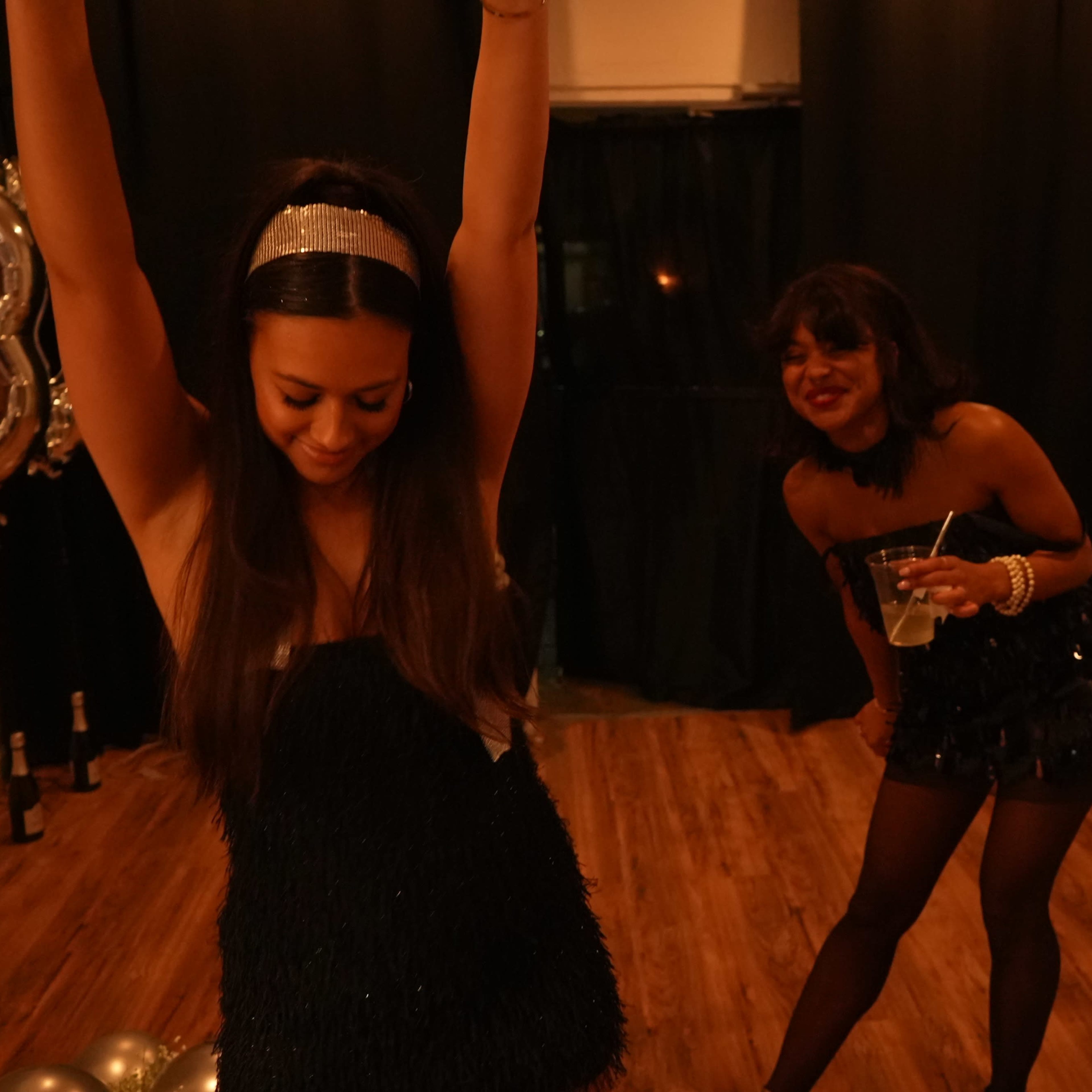 Three women are celebrating at a party, with one dancing enthusiastically while the others watch and take photos.