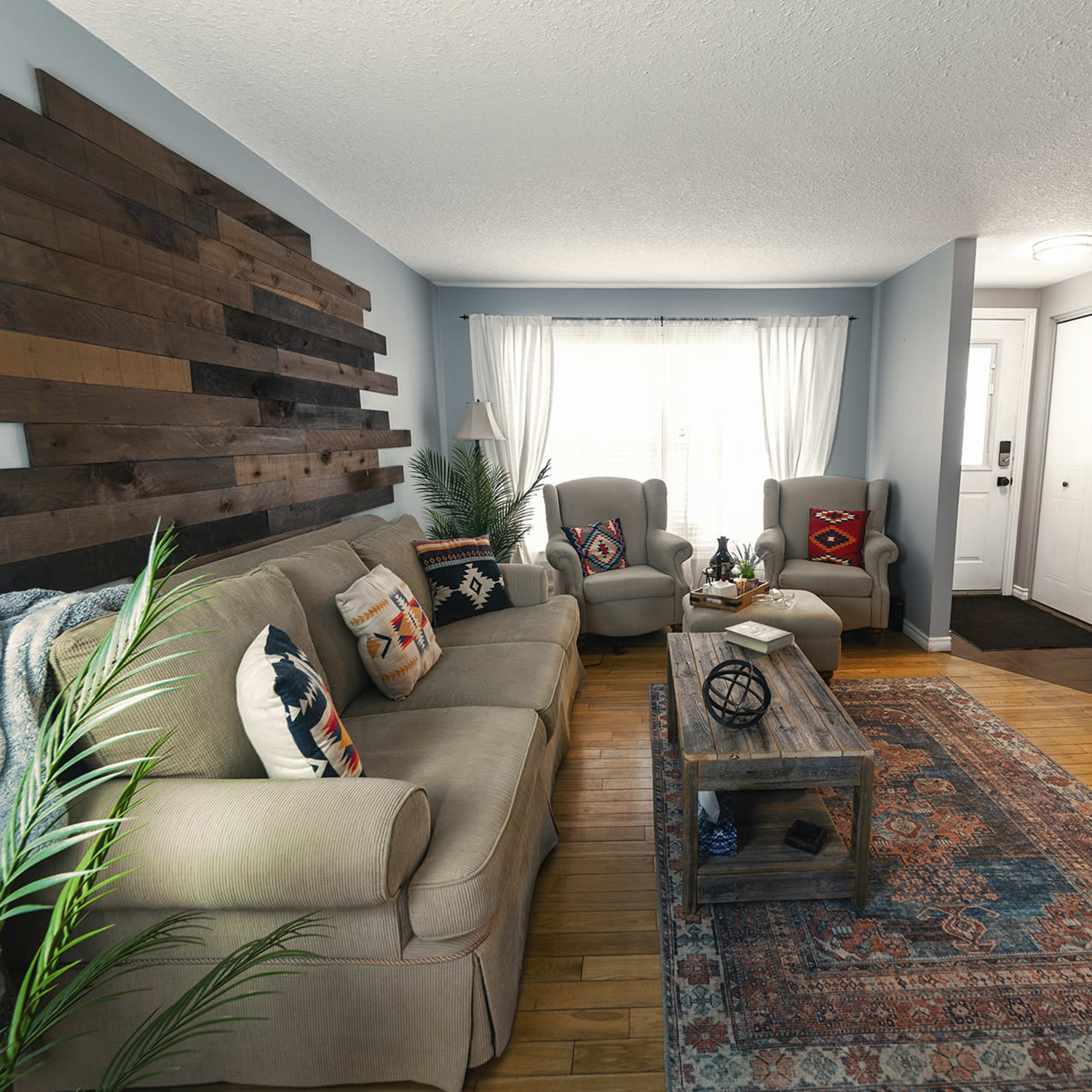 A living room features a wooden accent wall, a beige couch with decorative pillows, two armchairs, a coffee table, and a patterned area rug.