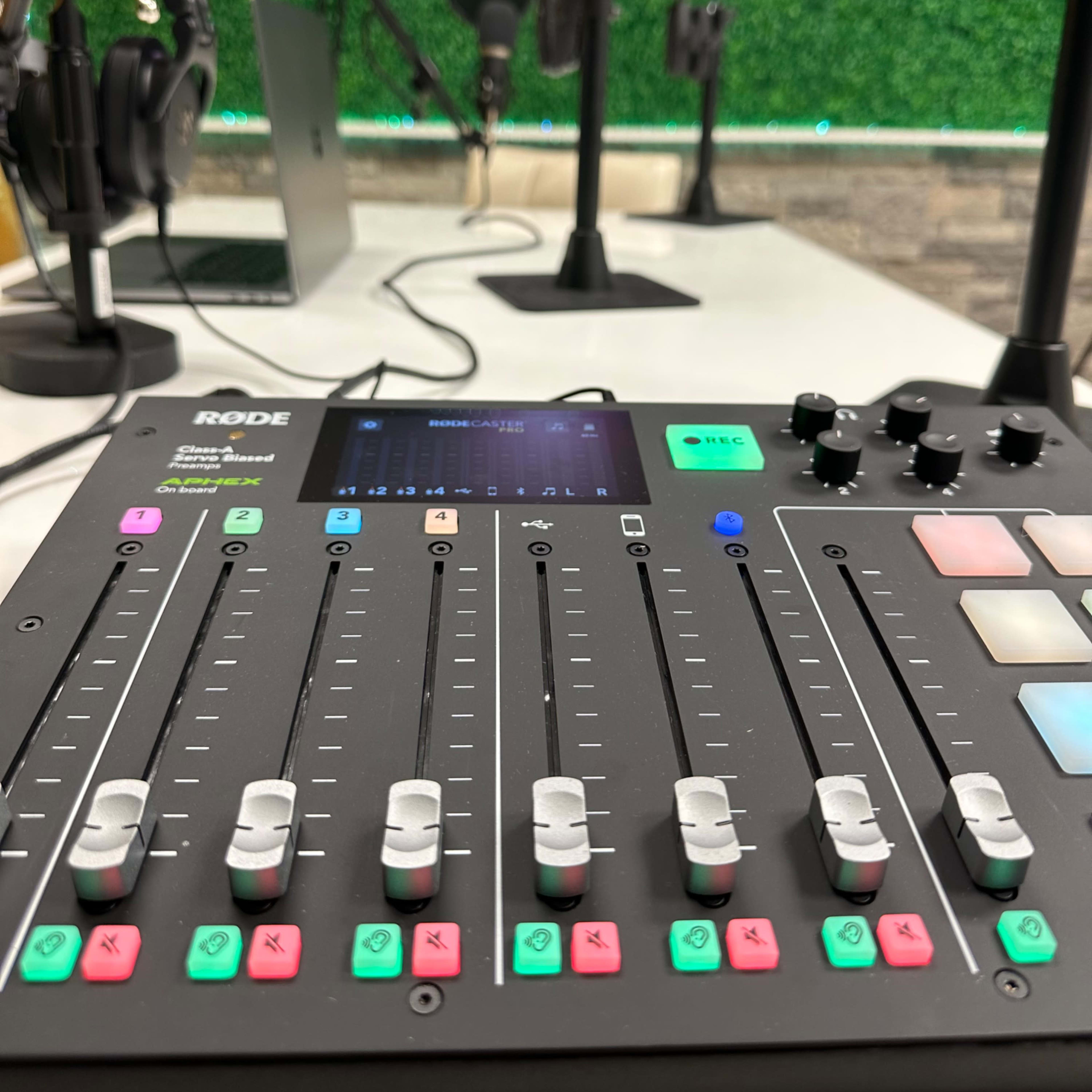 A close-up view of a Rode audio mixer with sliders and buttons, placed on a table in a recording studio featuring microphones and headphones in the background.