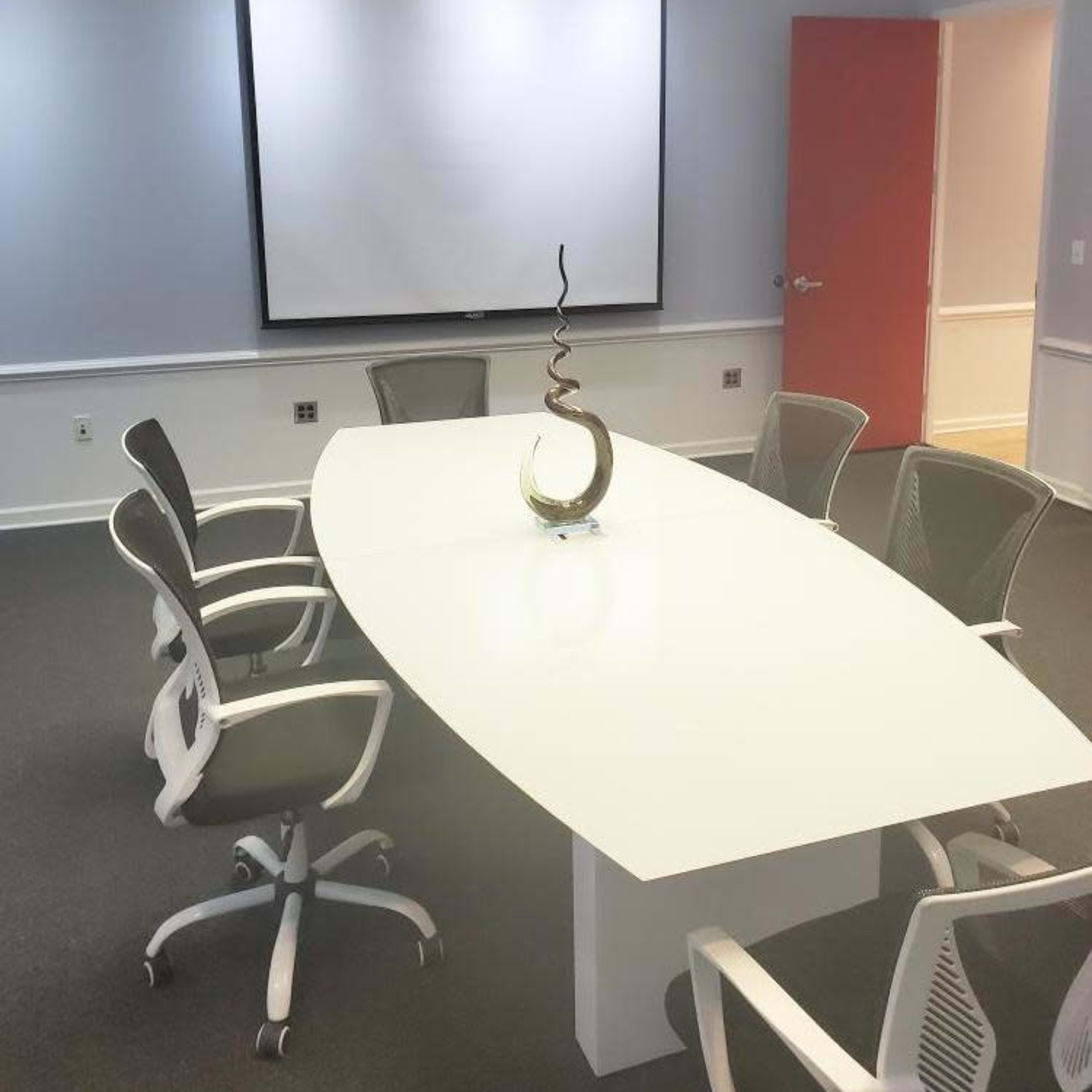 A modern conference room with a white oval table, five ergonomic chairs, and a decorative sculpture in the center, along with a large projection screen on the wall.