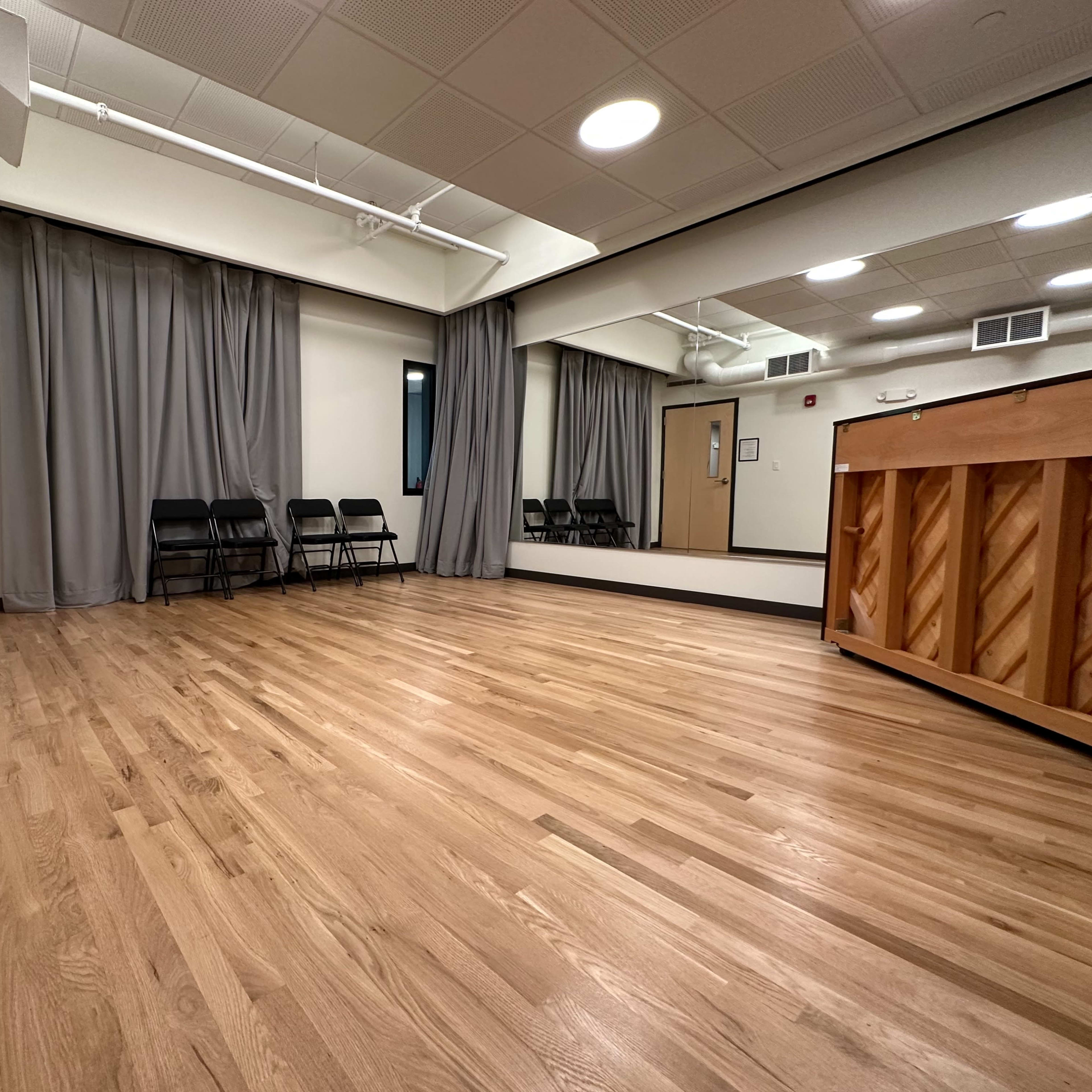 A spacious room with wooden flooring, gray curtains, a mirror, and several black chairs lined up along one wall.