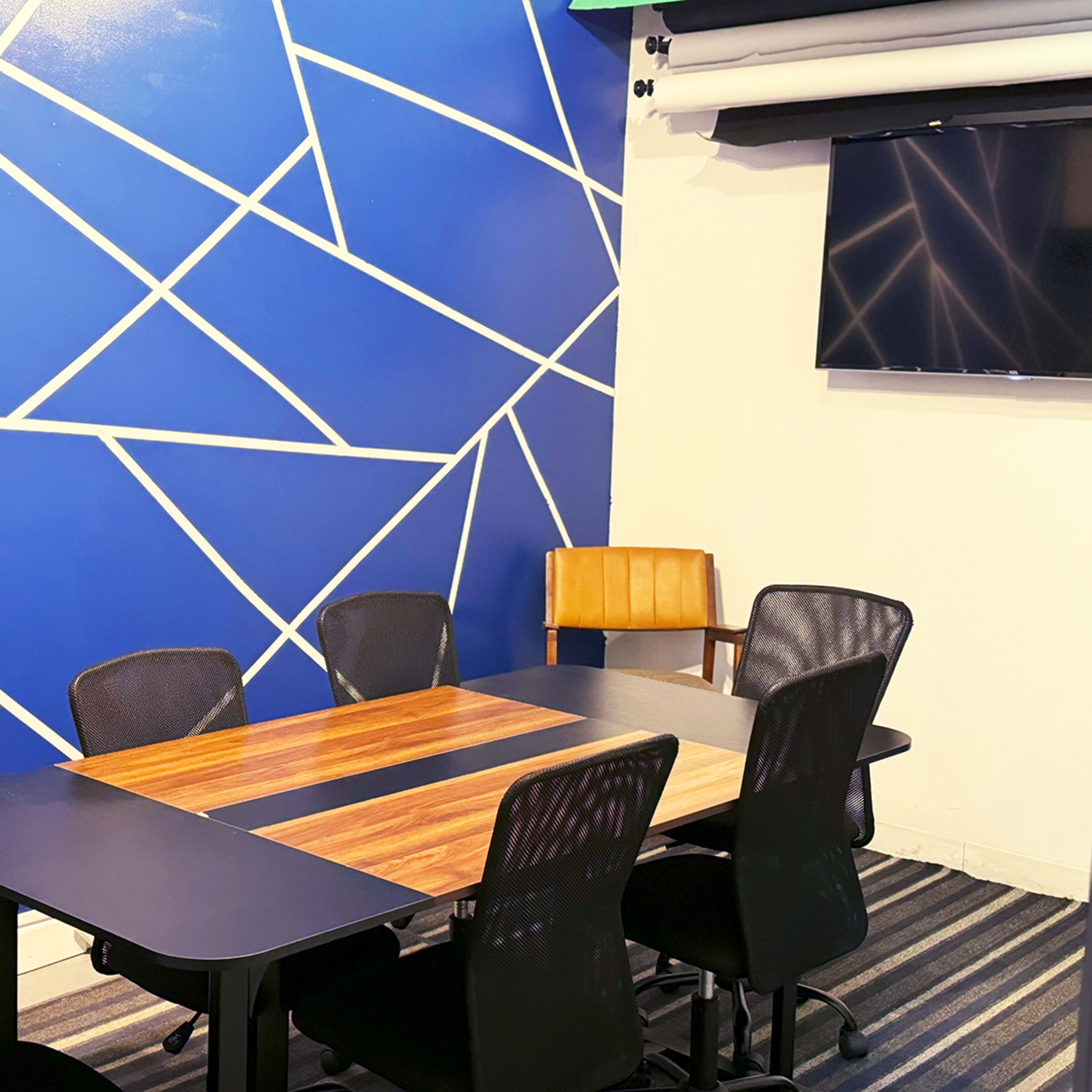A conference room features a wooden table surrounded by six black mesh chairs, with a blue geometric wall design and a mounted television.