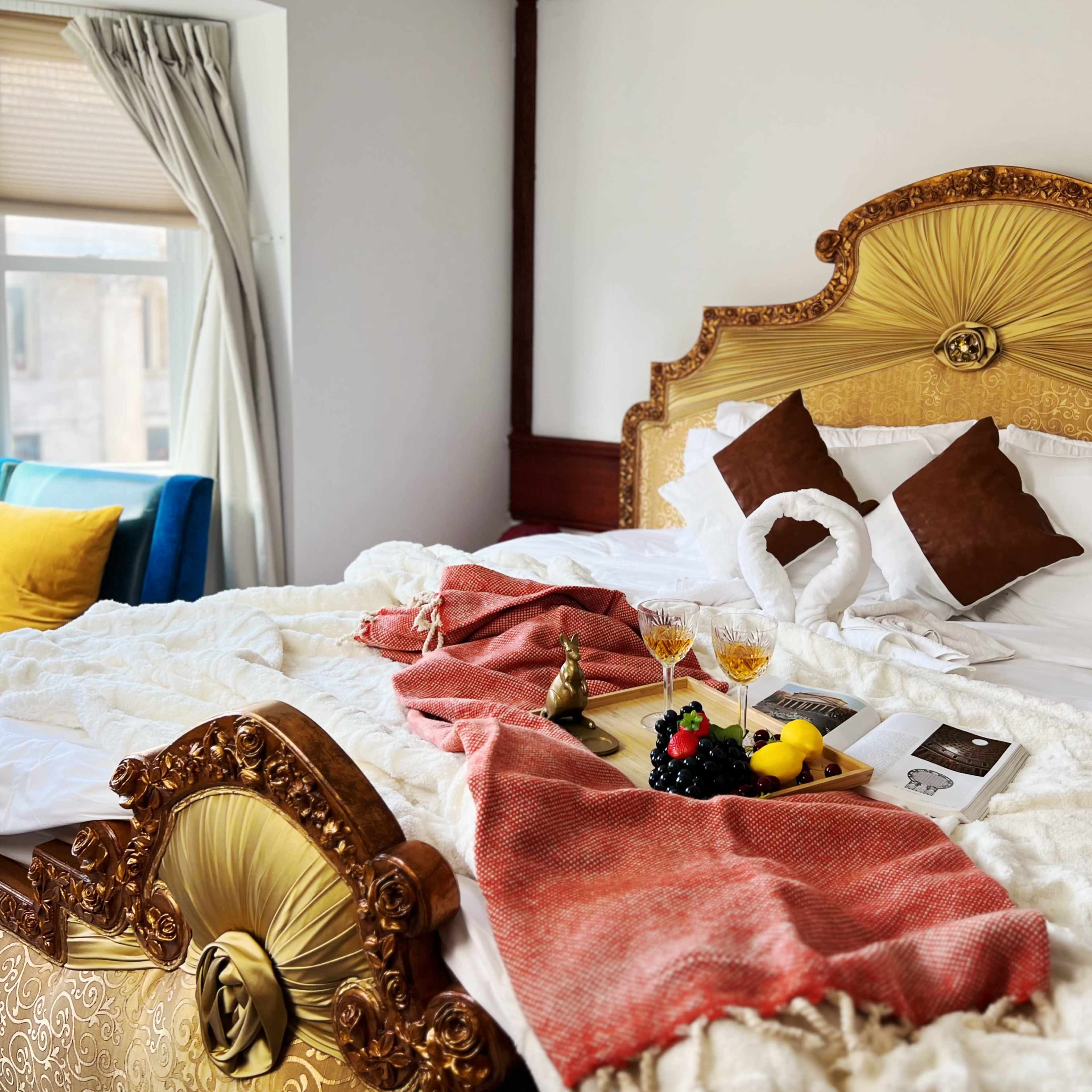A beautifully arranged bed with decorative pillows, a bottle of champagne, and a tray of fruits sits in a well-lit hotel room.