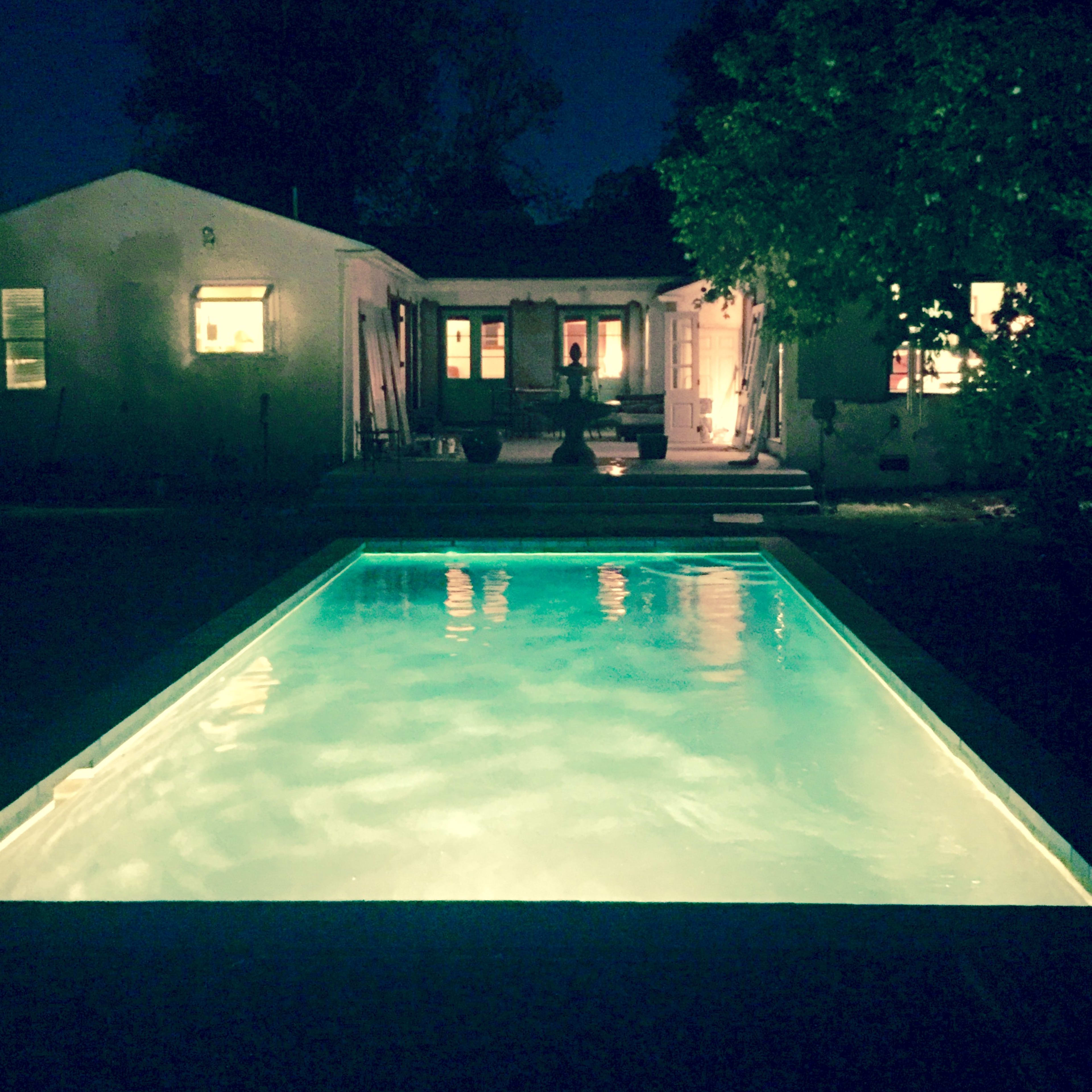 A well-lit swimming pool is positioned in front of a house at night, surrounded by trees and illuminated windows.