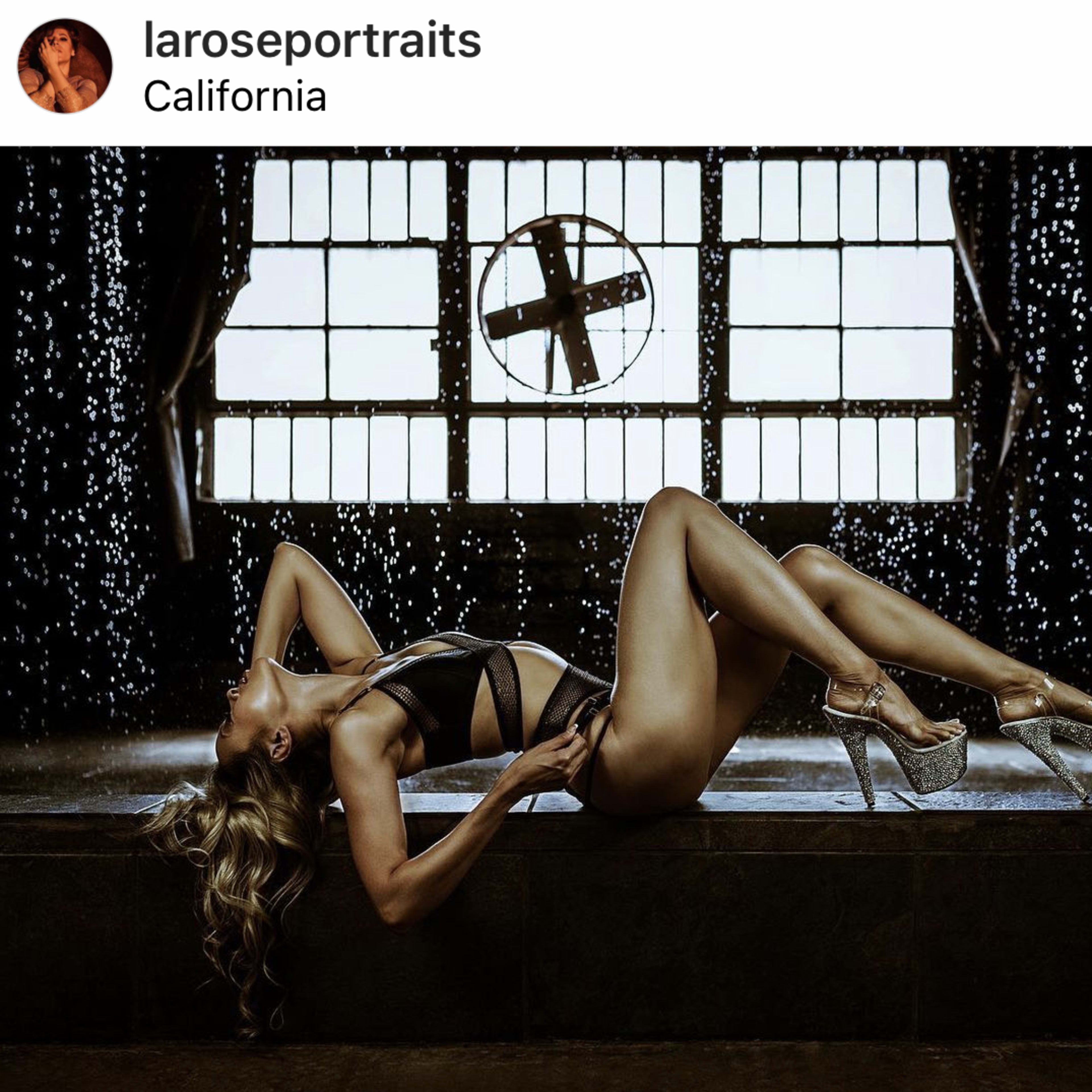 A model poses on her side in high heels in front of large windows with a cross-shaped design, while water cascades in the background.