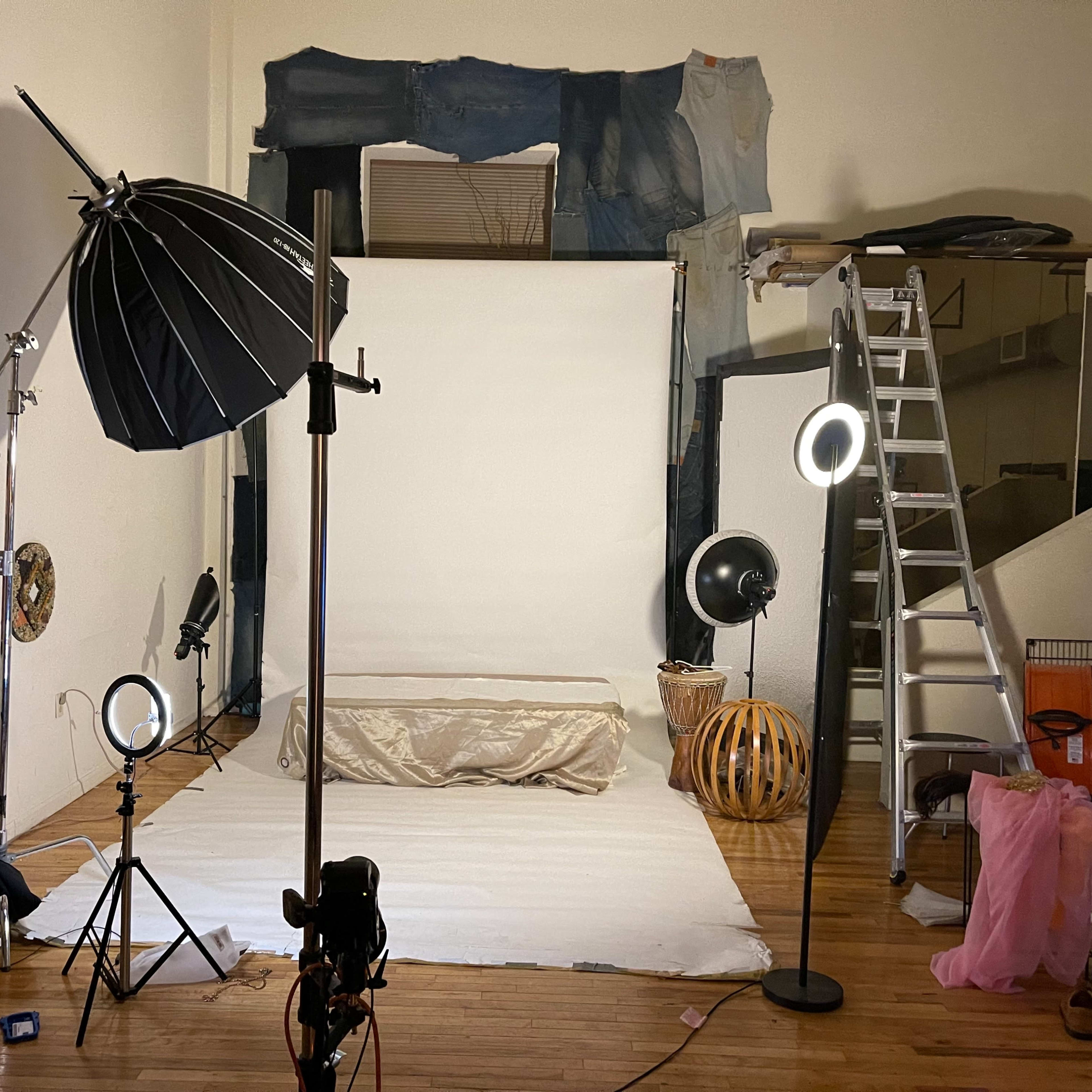 A photo studio setup with a white backdrop, lighting equipment, a ladder, and various props scattered around the floor.