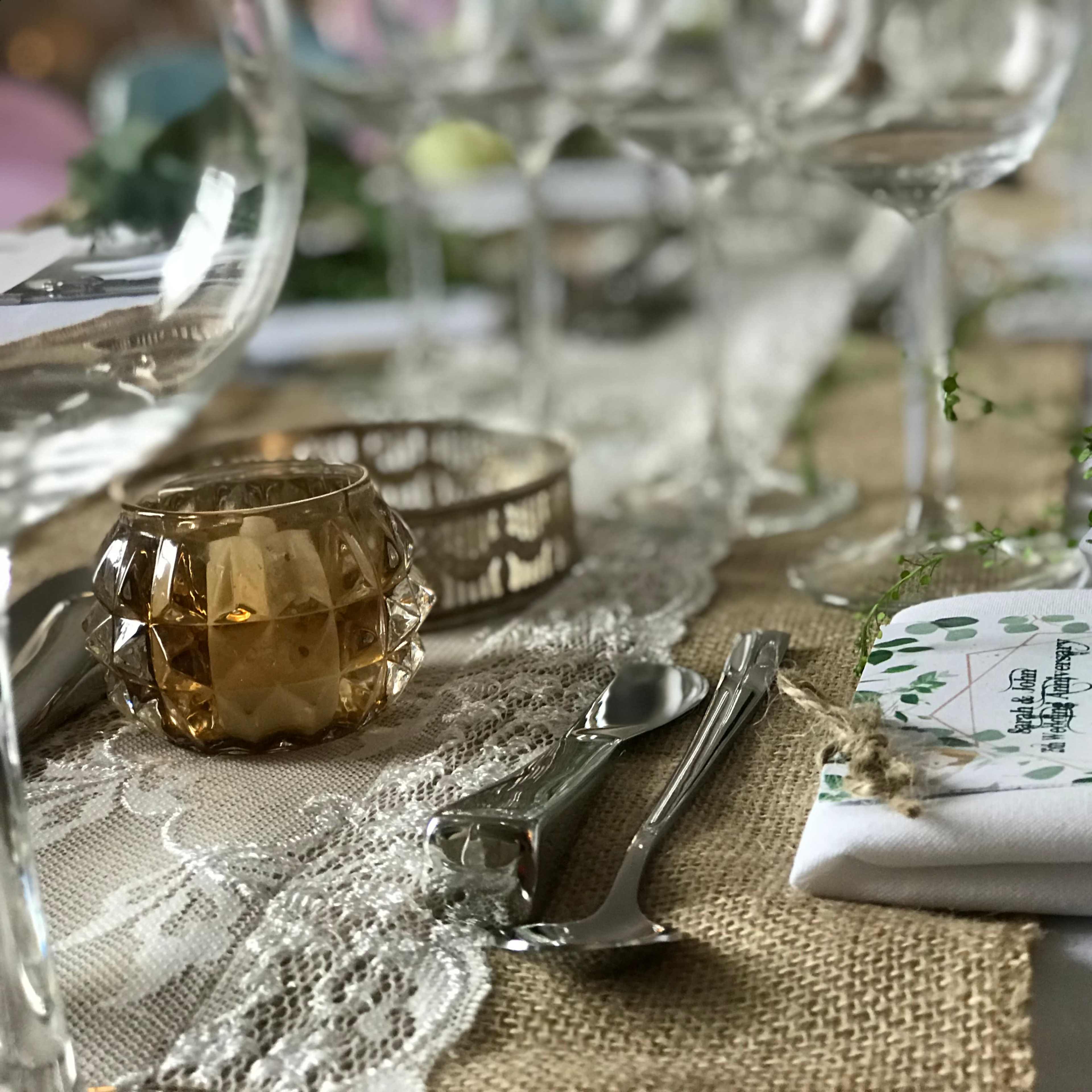 A neatly set dining table features glassware, cutlery, a decorative candle, and floral arrangements on a burlap runner.