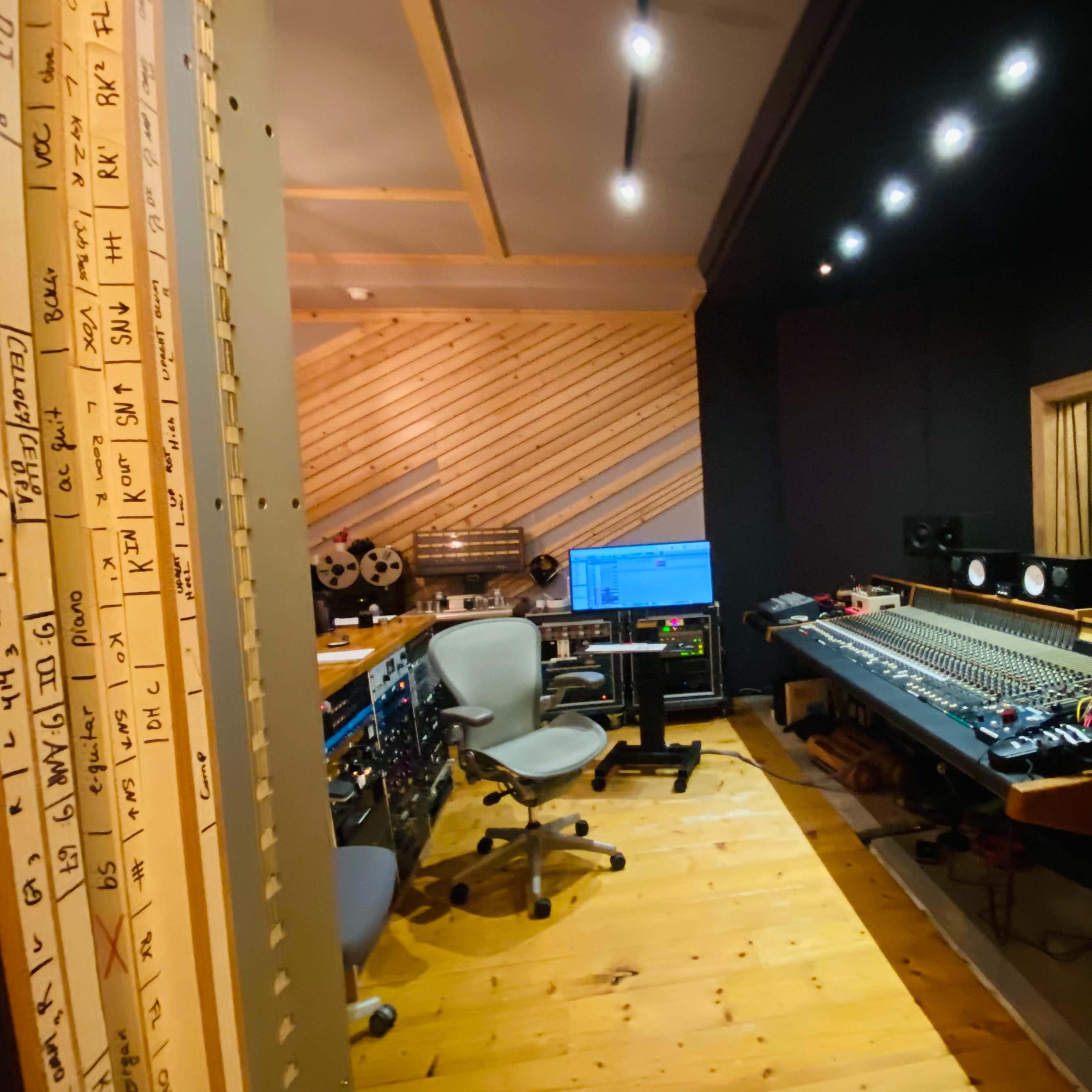 A recording studio with a mixing console, computer setup, and various audio equipment along wooden walls and a wooden floor.