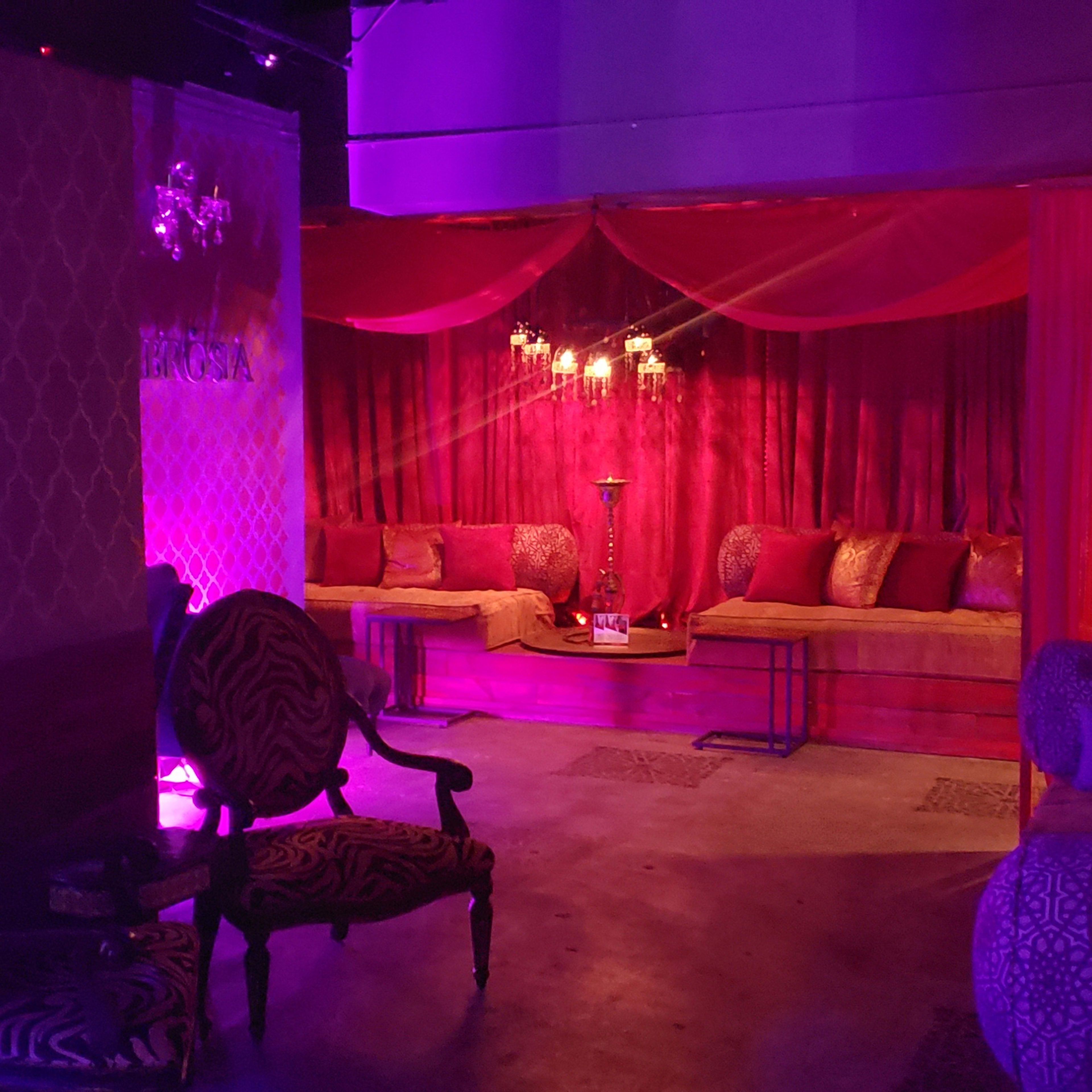 An interior space with plush couches, patterned chairs, and purple lighting, separated by sheer curtains.