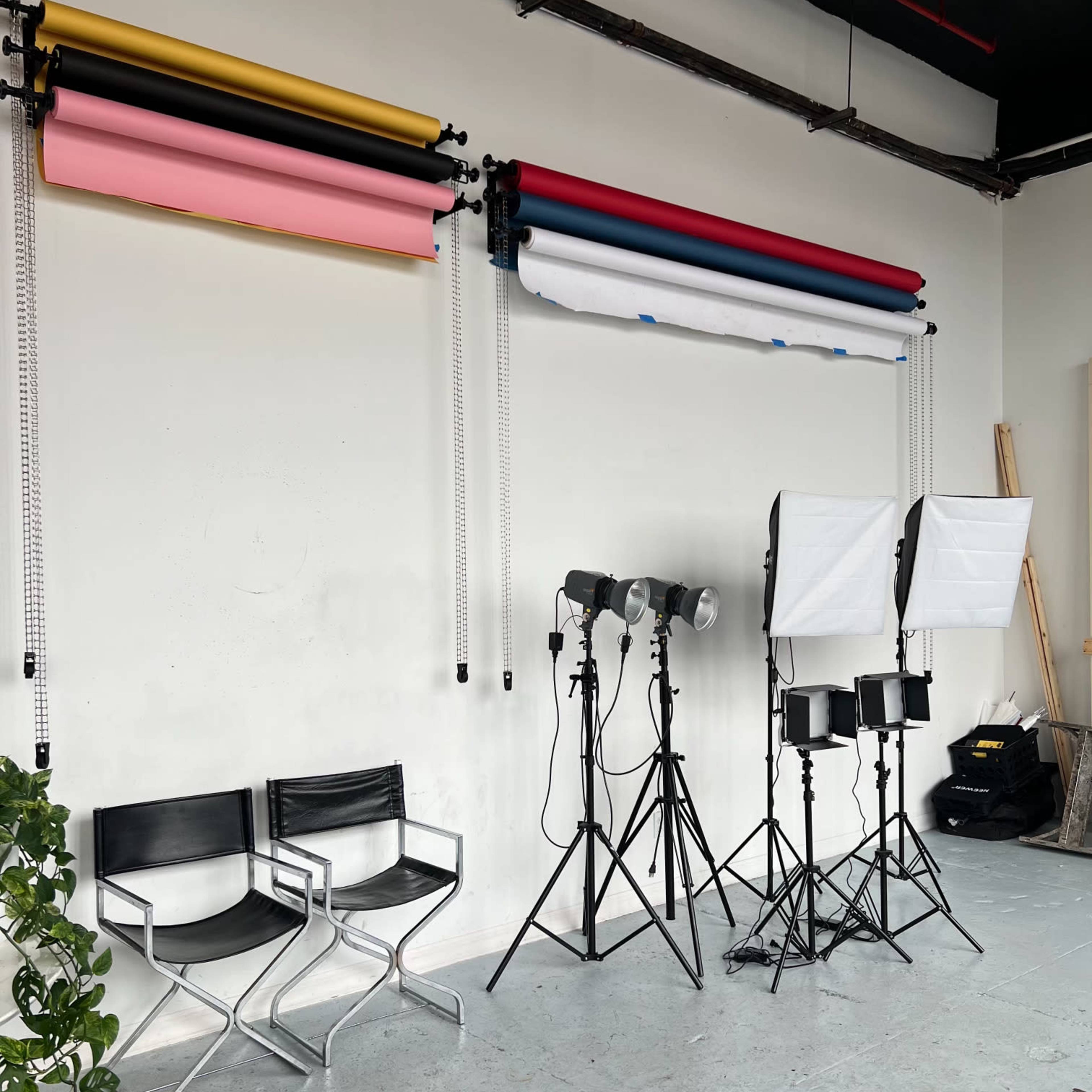 A photography studio setup featuring rolled backdrops in various colors, two black chairs, and multiple lighting stands arranged beside a wall.