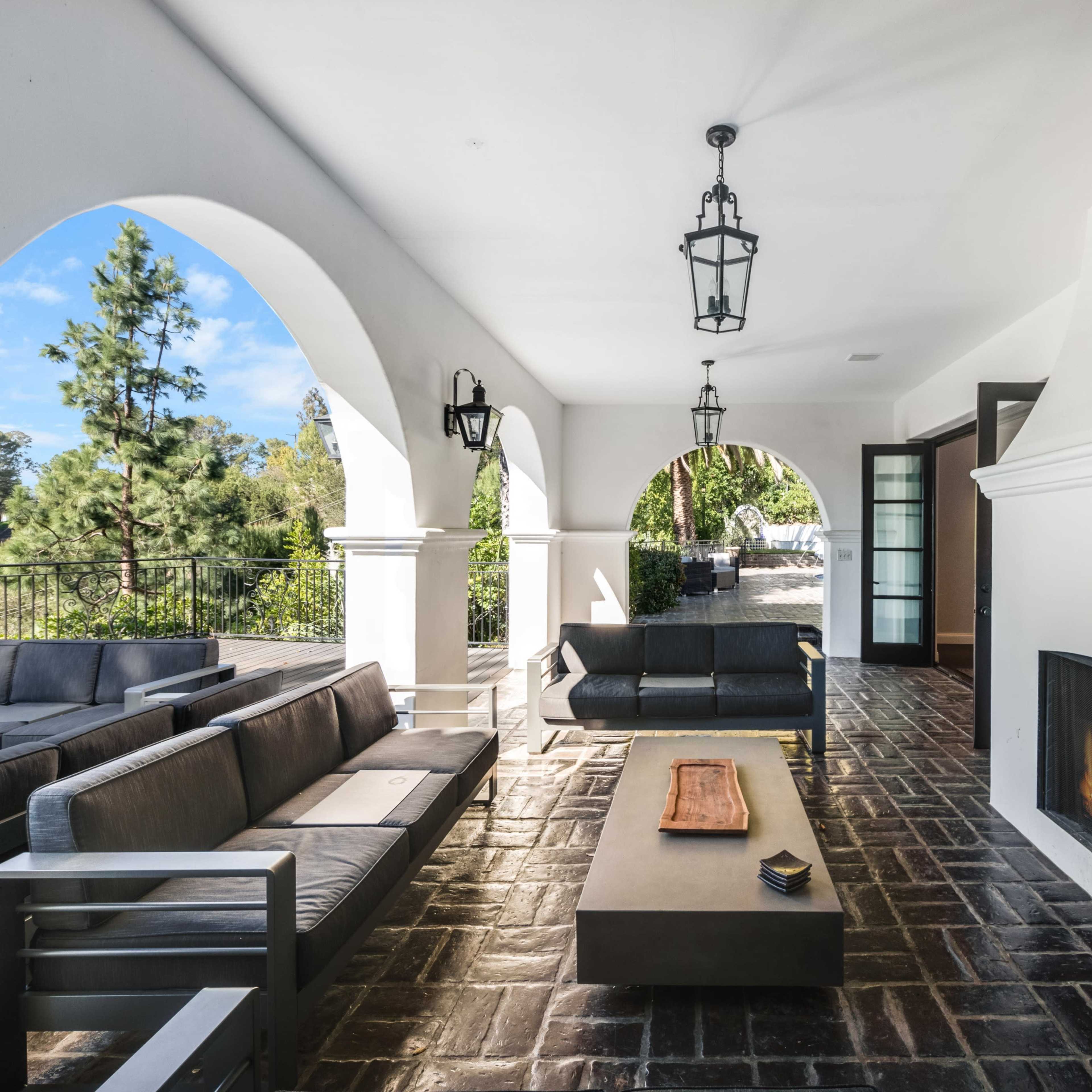 A covered outdoor living space featuring a modern seating arrangement, a coffee table, and a fireplace, with arches and large windows providing a view of the surrounding greenery.