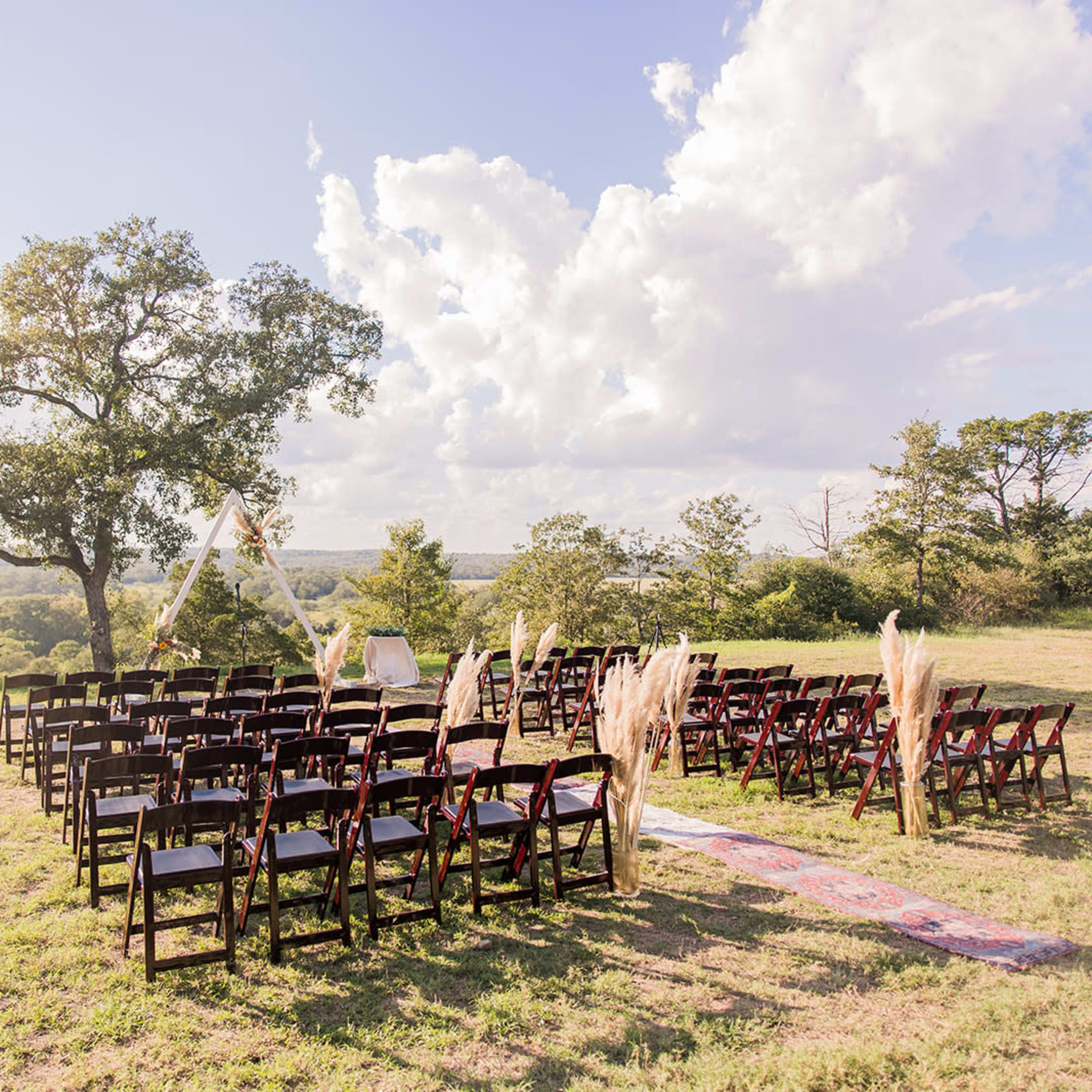 A wedding ceremony setup with rows of dark wooden chairs arranged on a grassy hillside under a partly cloudy sky.