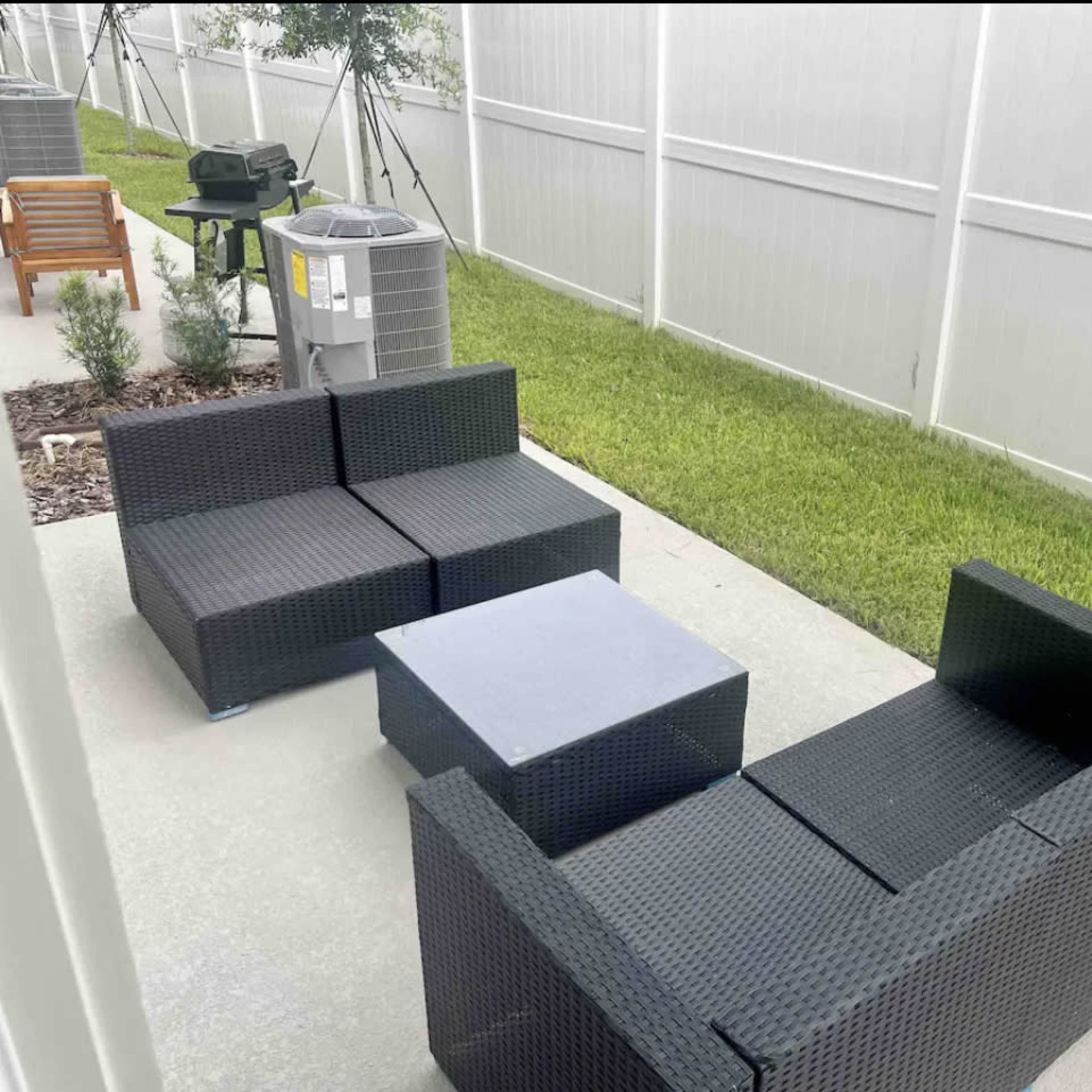 A small outdoor seating area with black wicker furniture, including a couch and a coffee table, set on a concrete patio next to a grassy area and a fence.