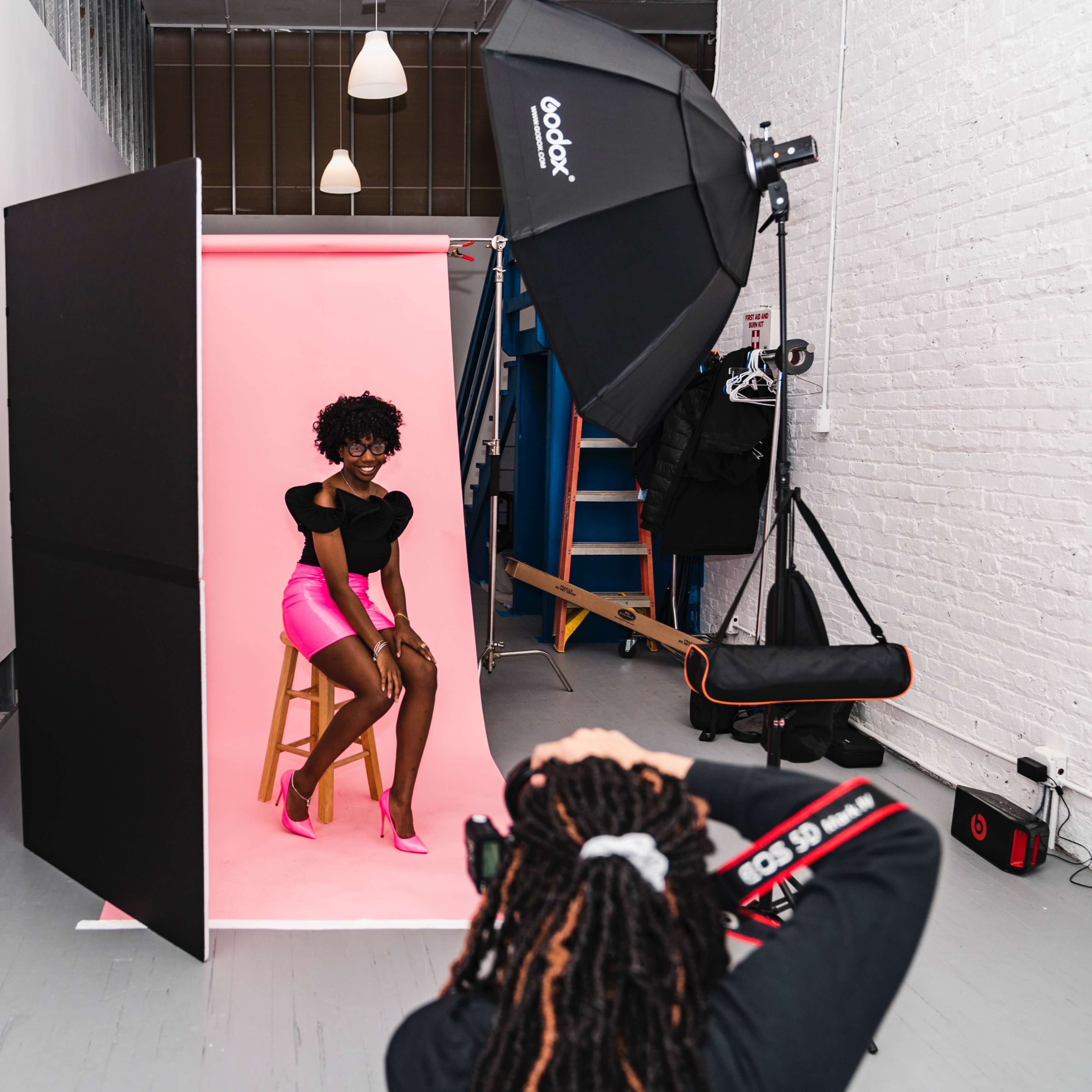 A model poses on a stool in front of a pink backdrop while a photographer captures the shot in a studio with various lighting equipment.