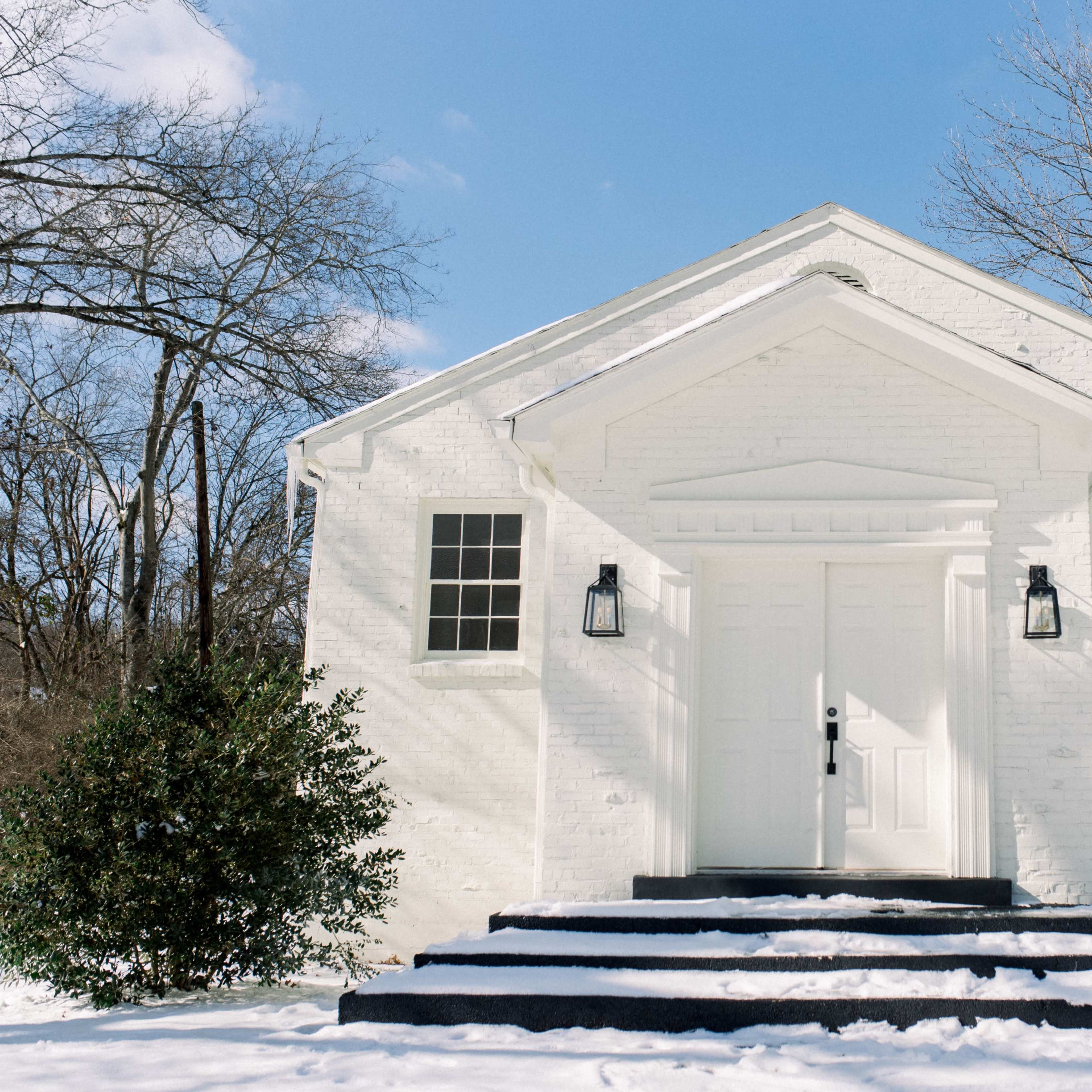 A white brick building with a gabled roof and two front windows stands in a snow-covered landscape.