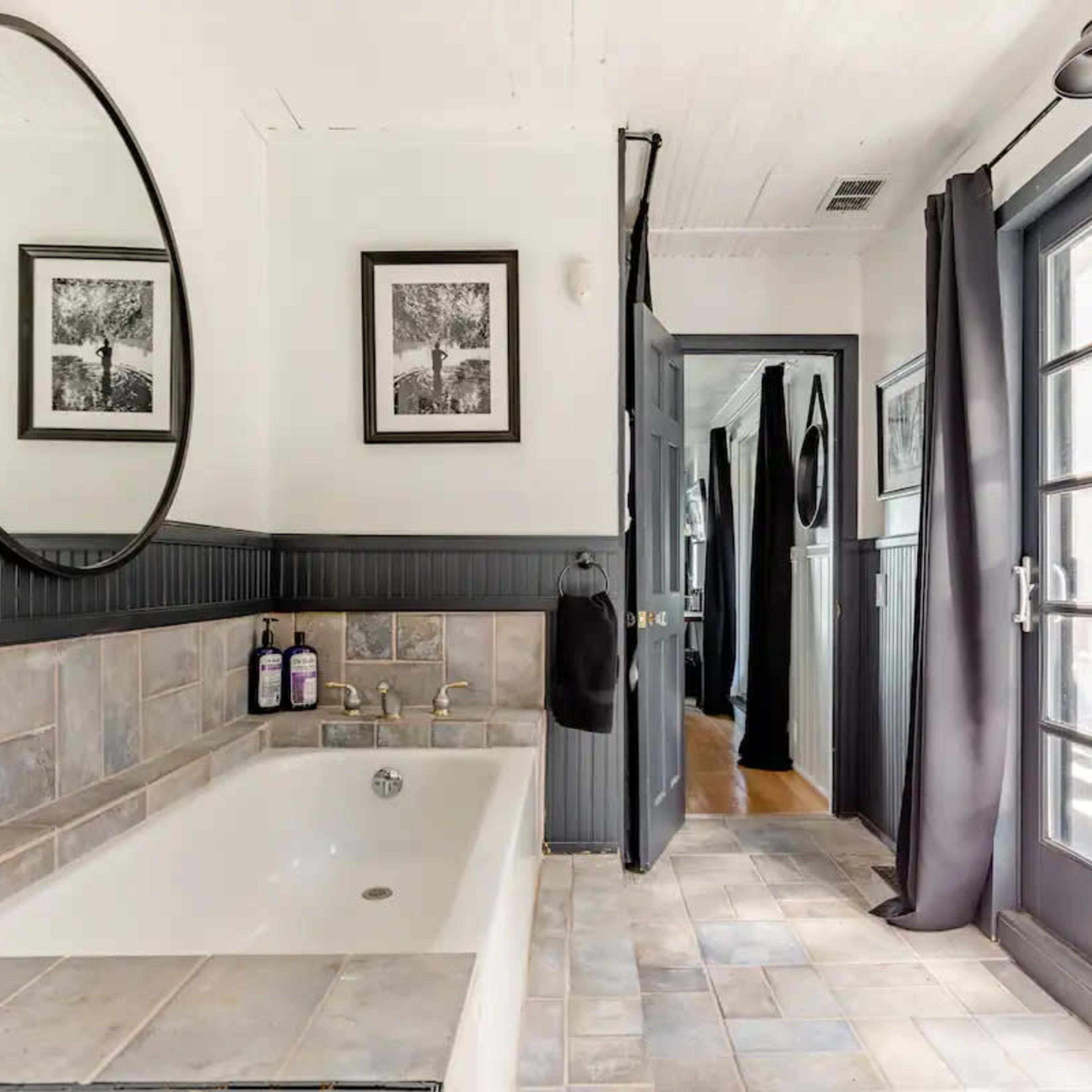 The bathroom features a freestanding tub, a round mirror, and framed photographs on the wall, with dark curtains and double doors leading outside.