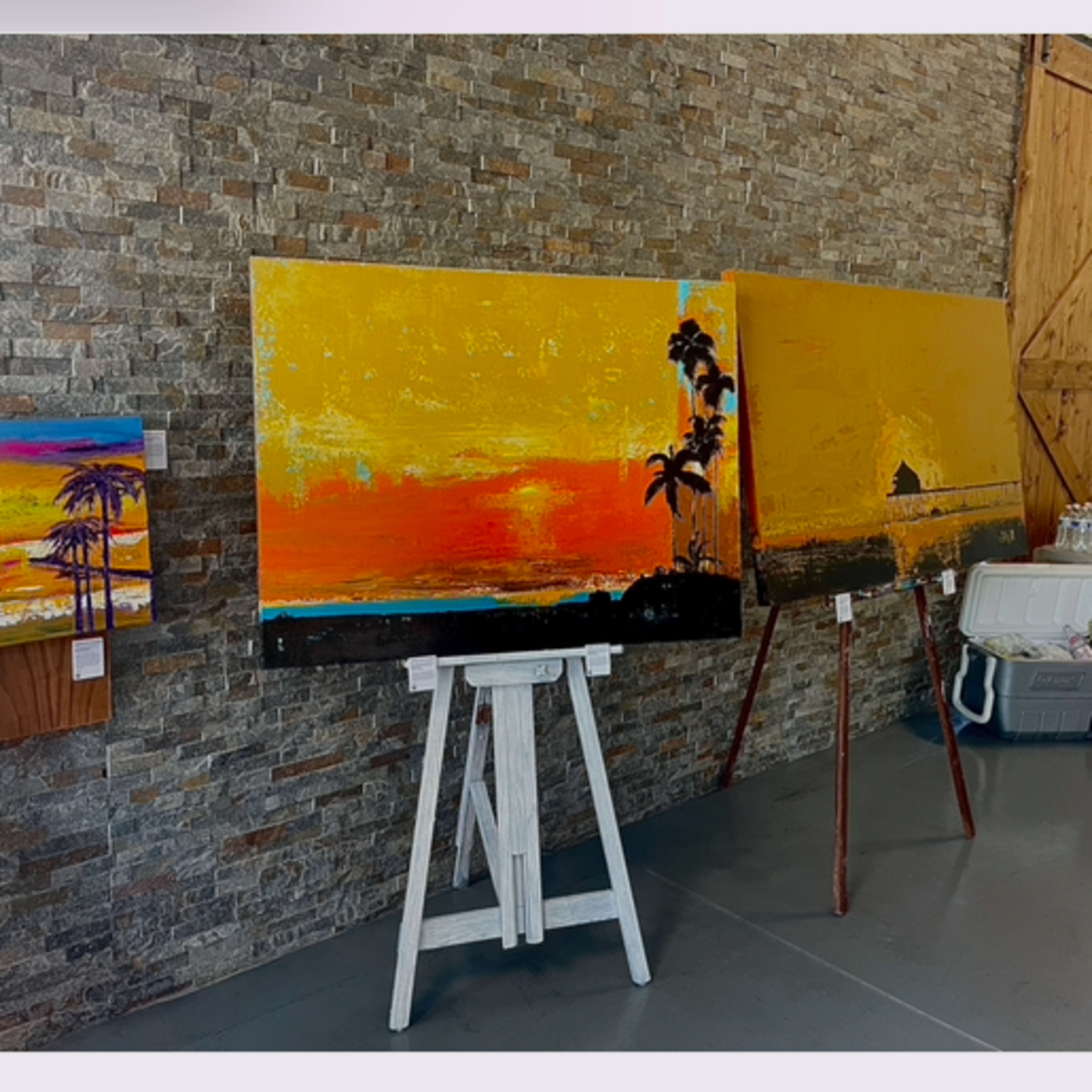 An art gallery with two distinct areas: one features a large painting of a sunset on an easel against a stone wall, while the other displays various framed artworks on a white wall, with a person in colorful attire examining the pieces.