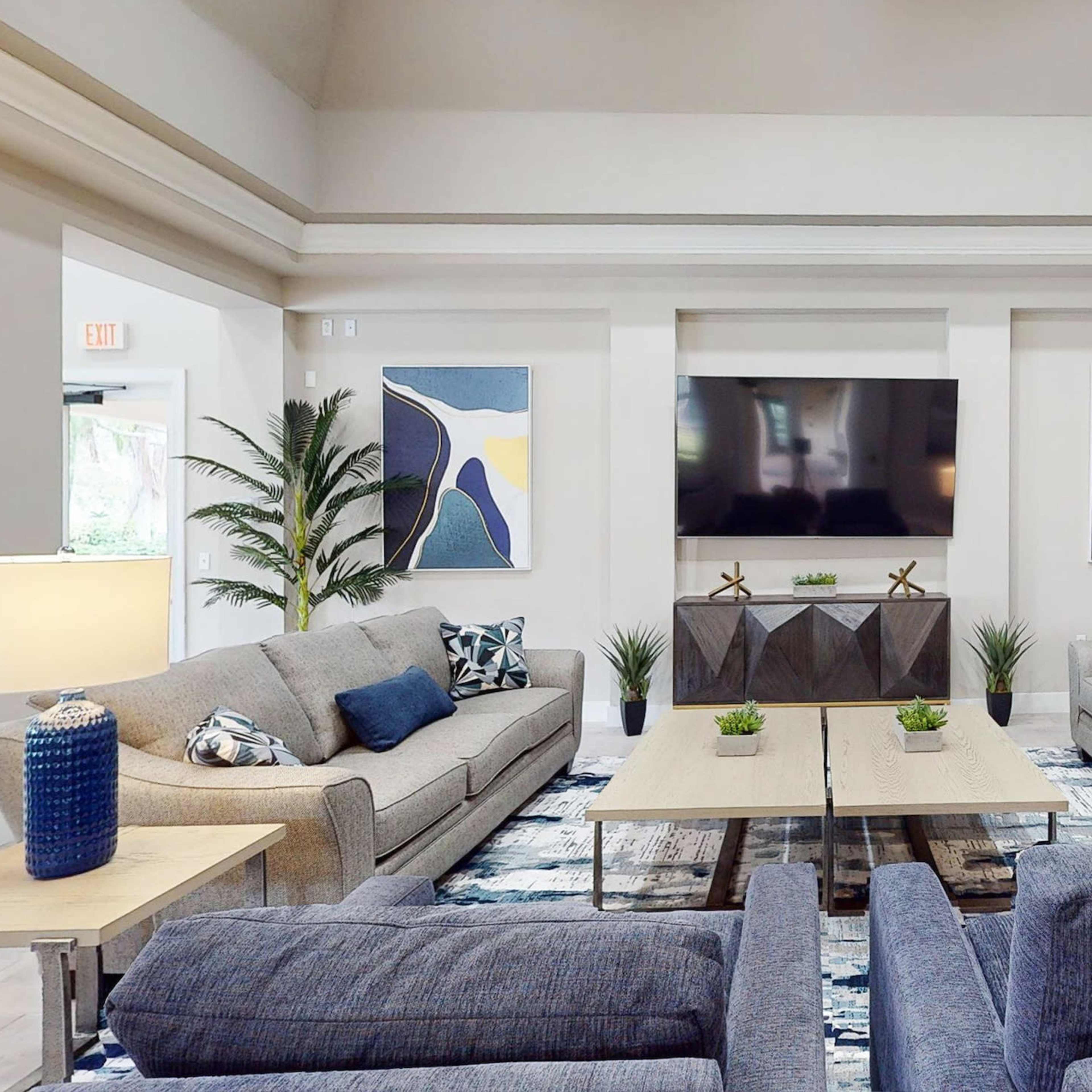 A spacious modern living area with two gray sofas and accent chairs arranged around coffee tables, featuring plants and abstract artwork on the walls.