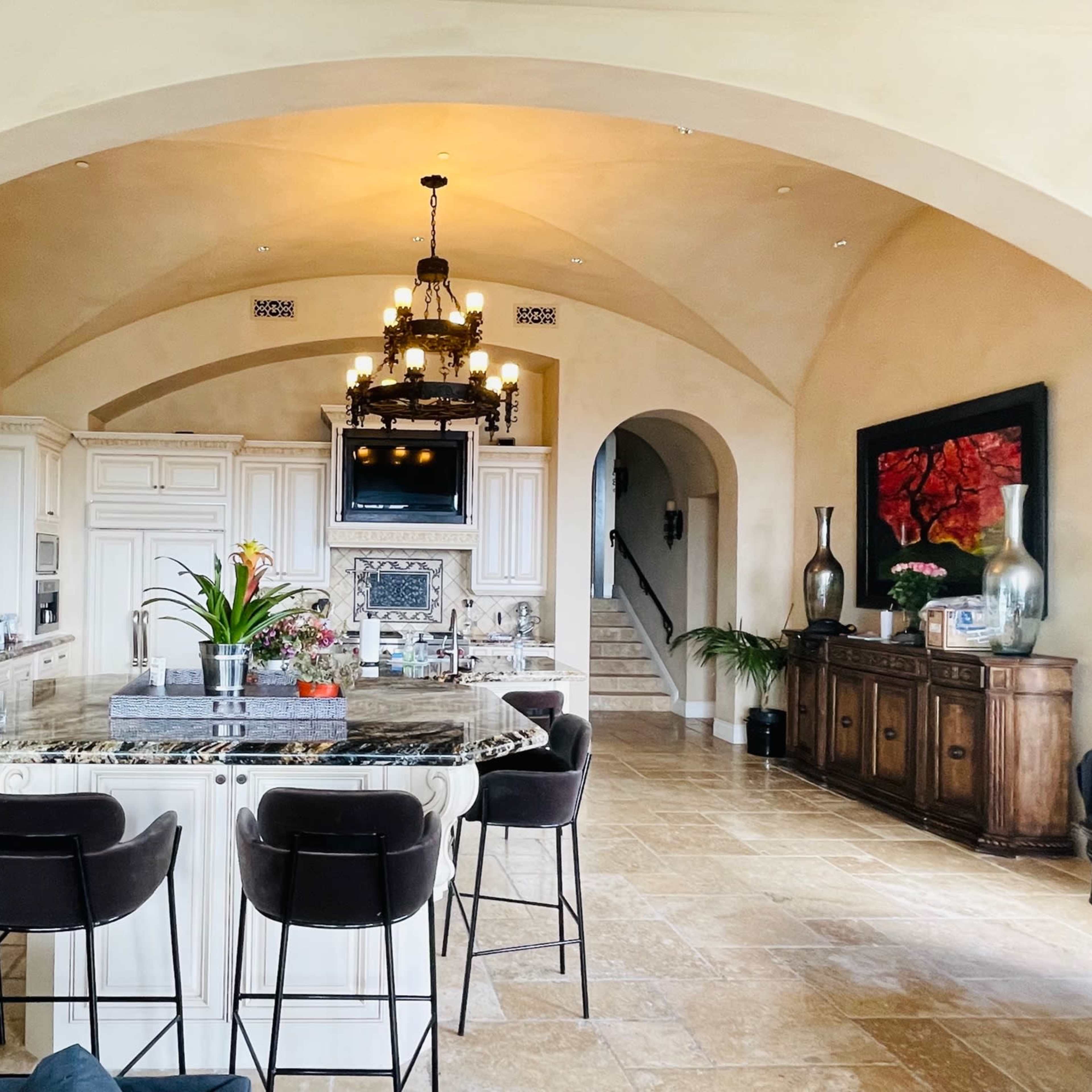 A spacious kitchen featuring a large island with bar stools, white cabinetry, and an arched ceiling, adjacent to a living area and staircase.