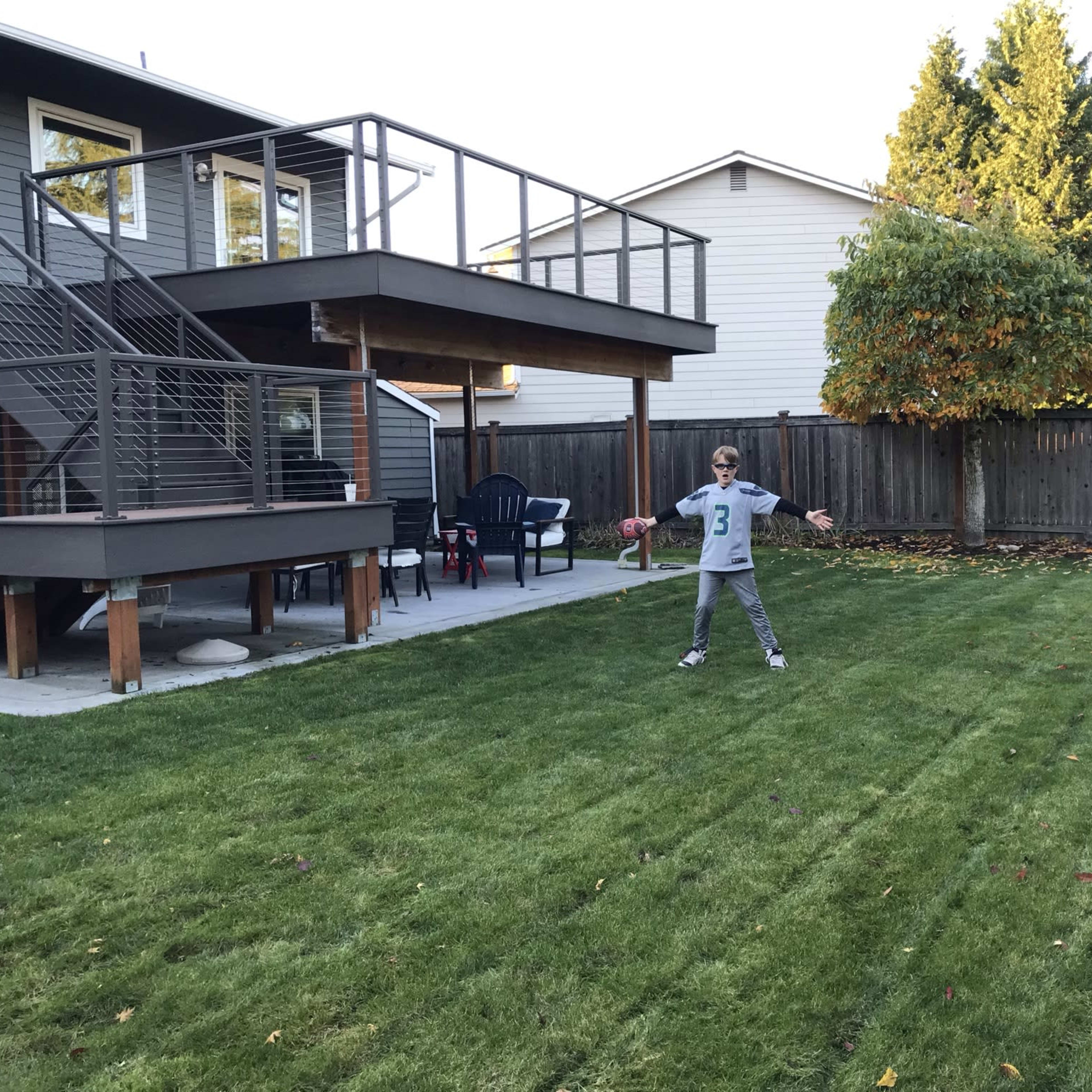A child stands in a neatly mowed backyard with a deck in the background and a few lawn chairs nearby.